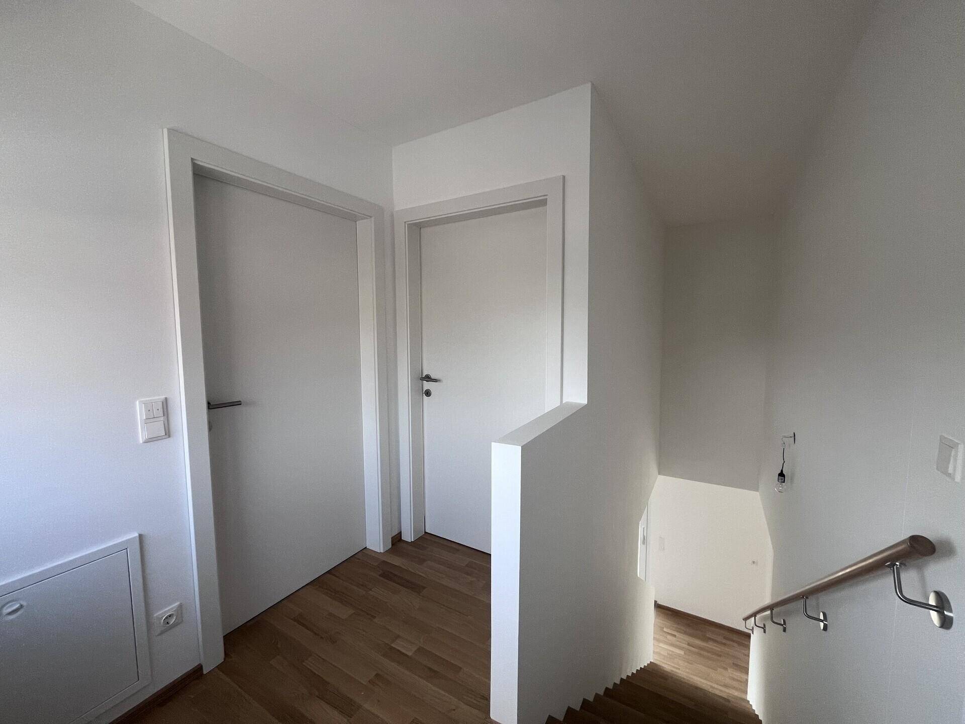 Heller Flur mit Holztreppe und weißen Türen, der die Stockwerke verbindet.