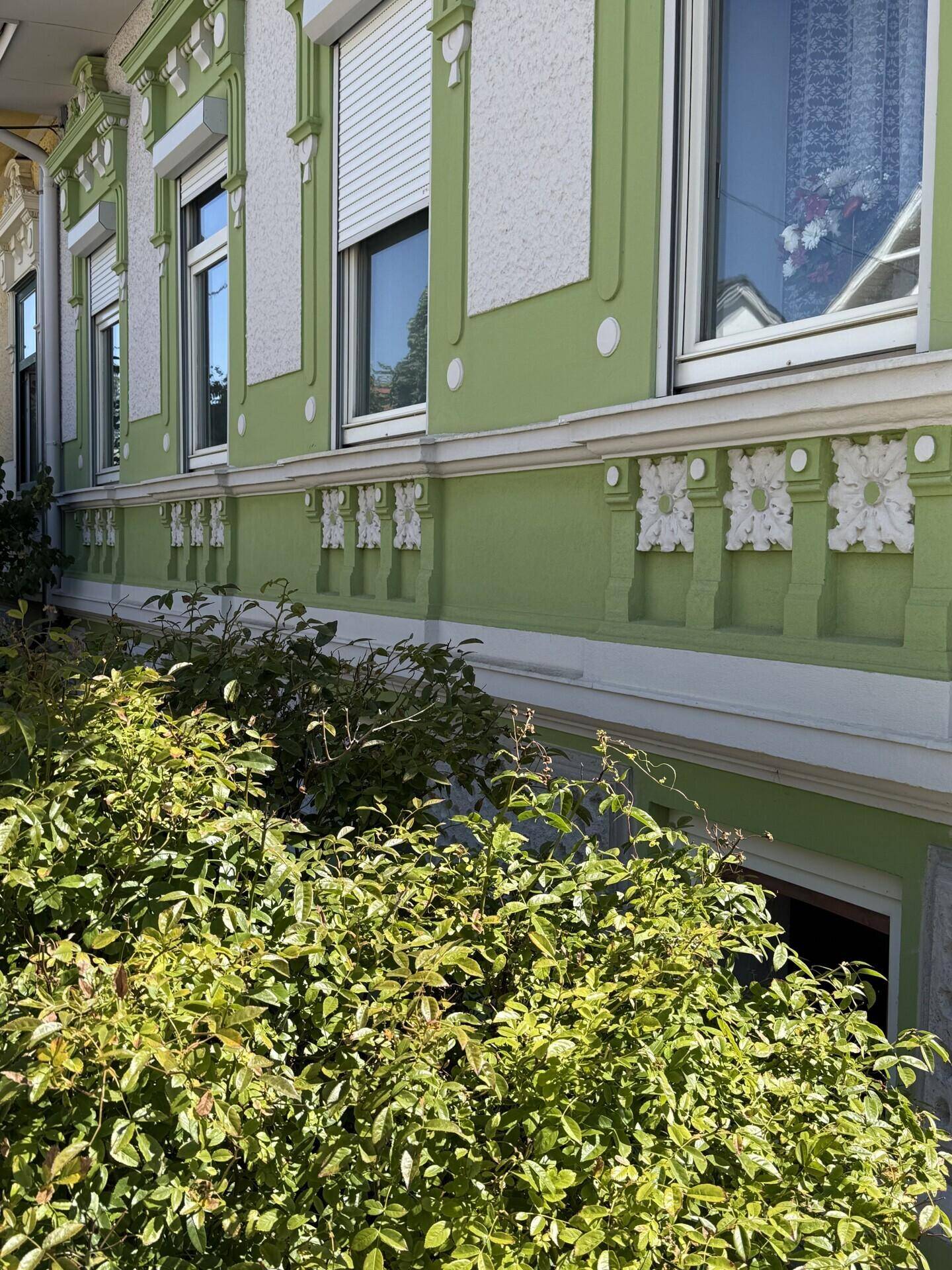 Detailansicht der grünen Fassade mit dekorativen Stuckelementen und Fenstern mit Rollläden.