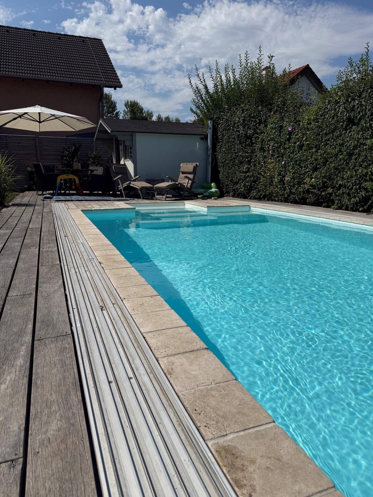 Großer Außenpool mit Holzdeck und Liegebereich, umgeben von einer grünen Hecke.