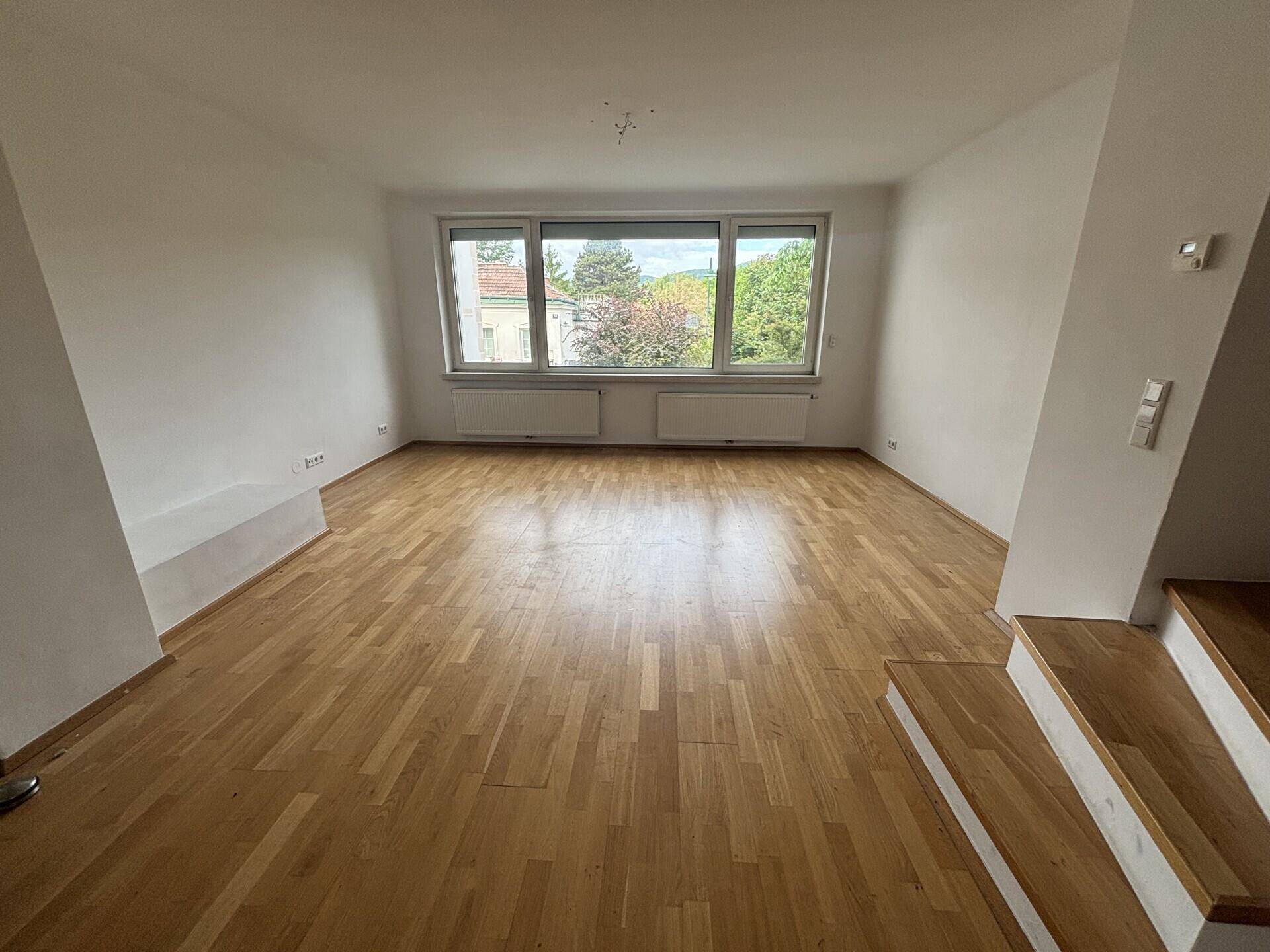 Helles, leeres Wohnzimmer mit Parkettboden und großen Fenstern mit Blick ins Grüne.