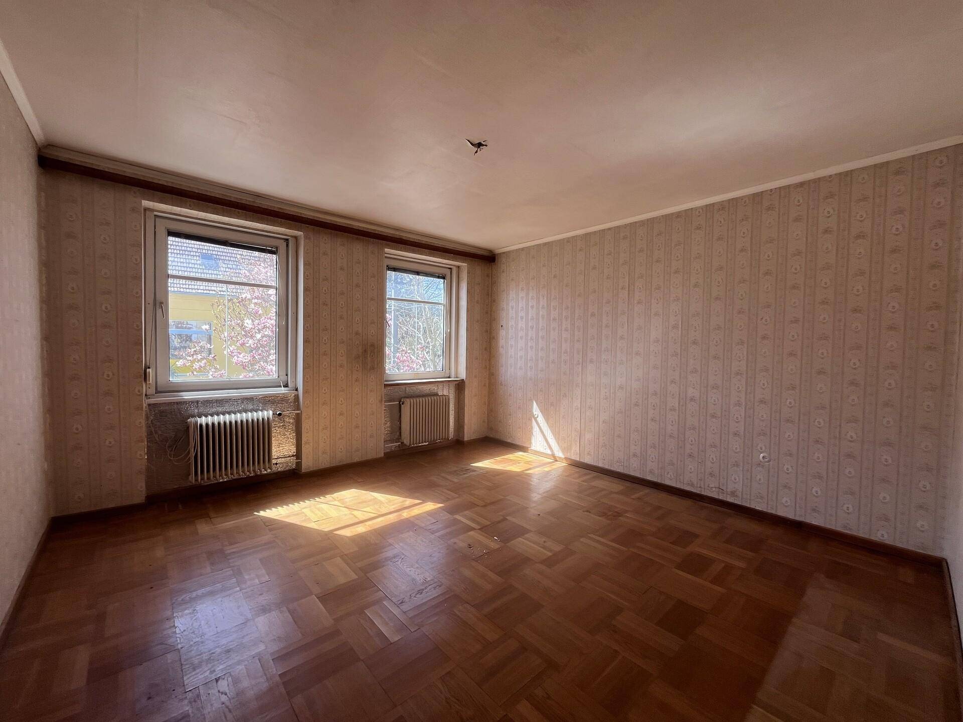 Geräumiges Zimmer mit Fischgrätparkett, zwei Fenstern und alter Tapete.