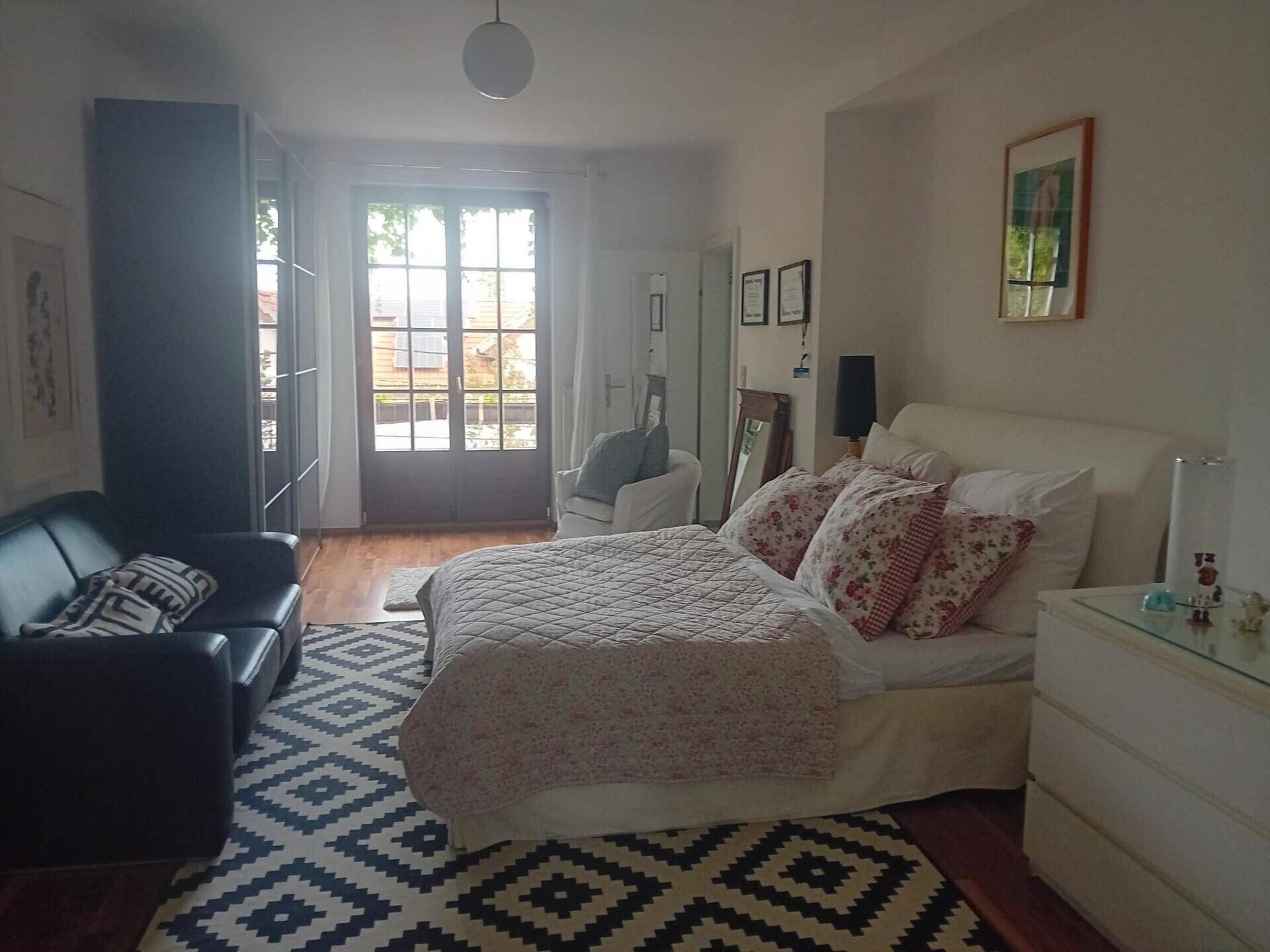 Helles Schlafzimmer mit Doppelbett, Sofa und großem Fenster, Holzboden.