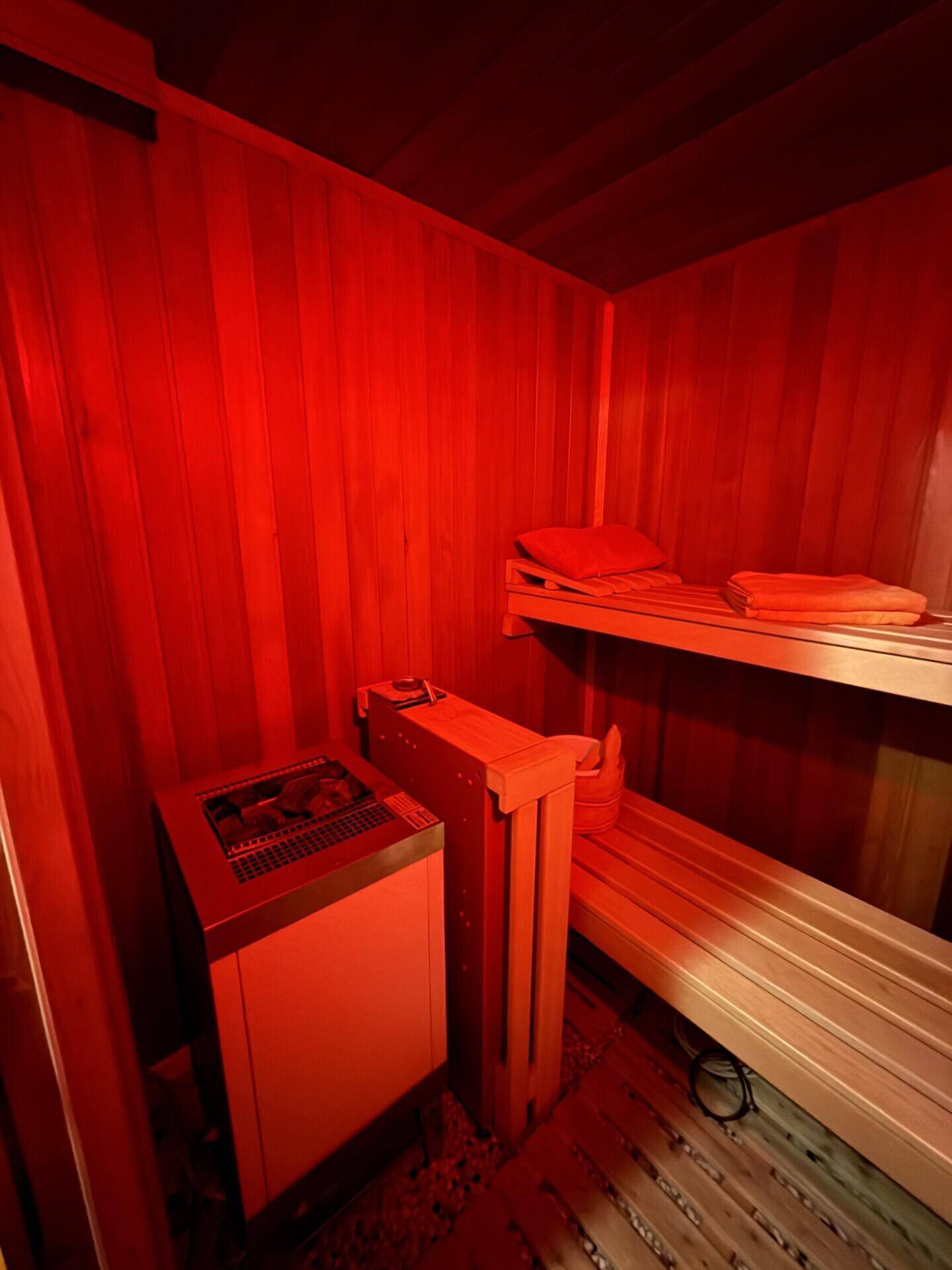 Gemütliche Sauna mit Holzbänken und Heizofen, beleuchtet in warmem Rotlicht.