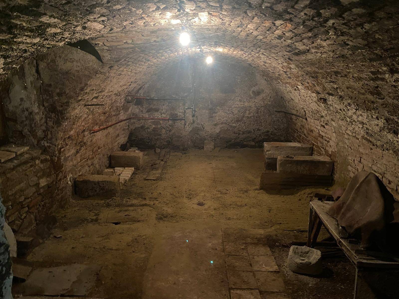 Historischer Gewölbekeller mit Ziegelwänden und sandigem Boden, beleuchtet von Glühbirnen.
