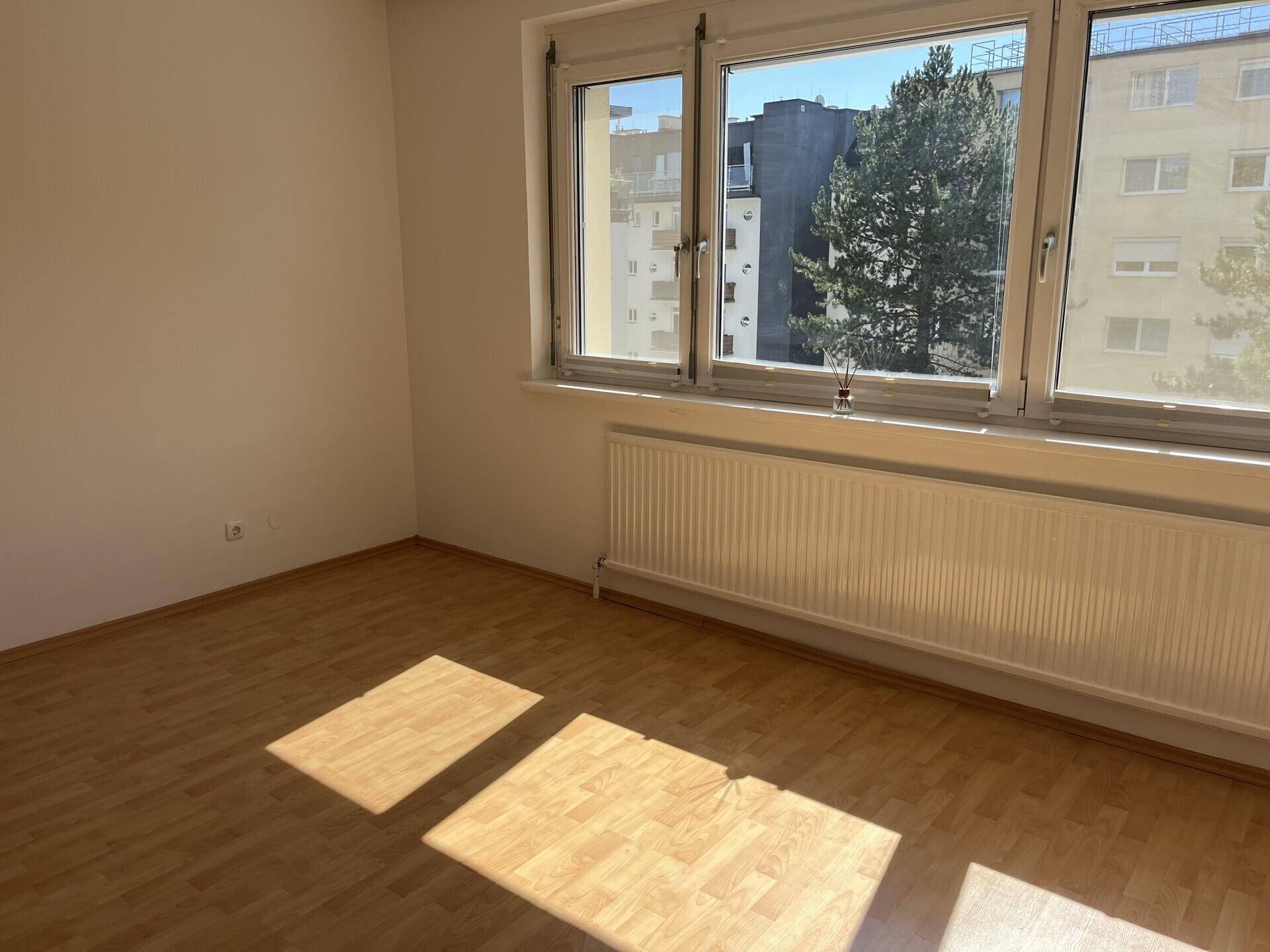 Sonniger Raum mit Laminatboden und Blick auf die umliegenden Gebäude und Bäume.