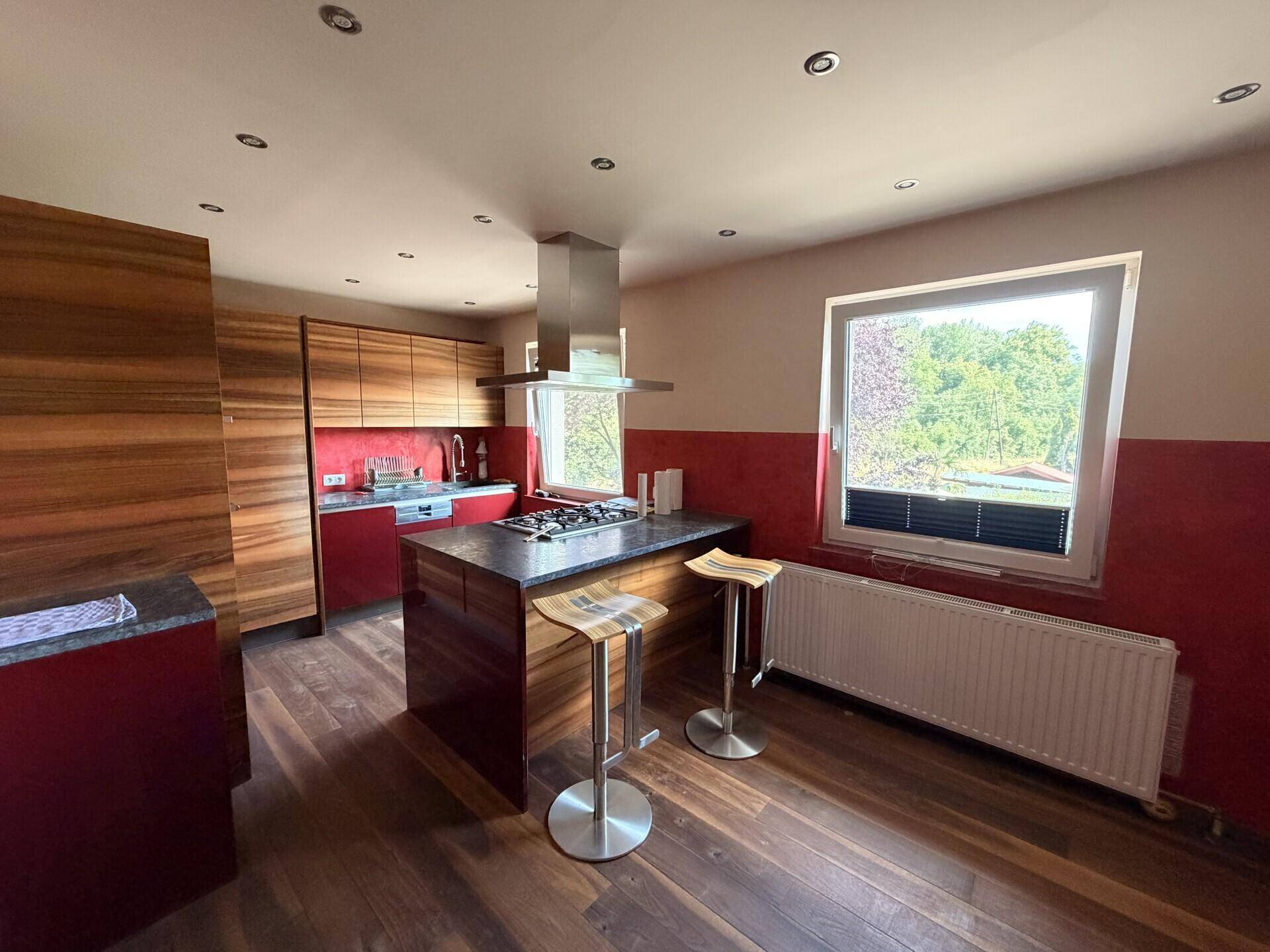 Moderne Küche mit Holzschränken, roter Akzentwand, Kochinsel und Blick ins Grüne.
