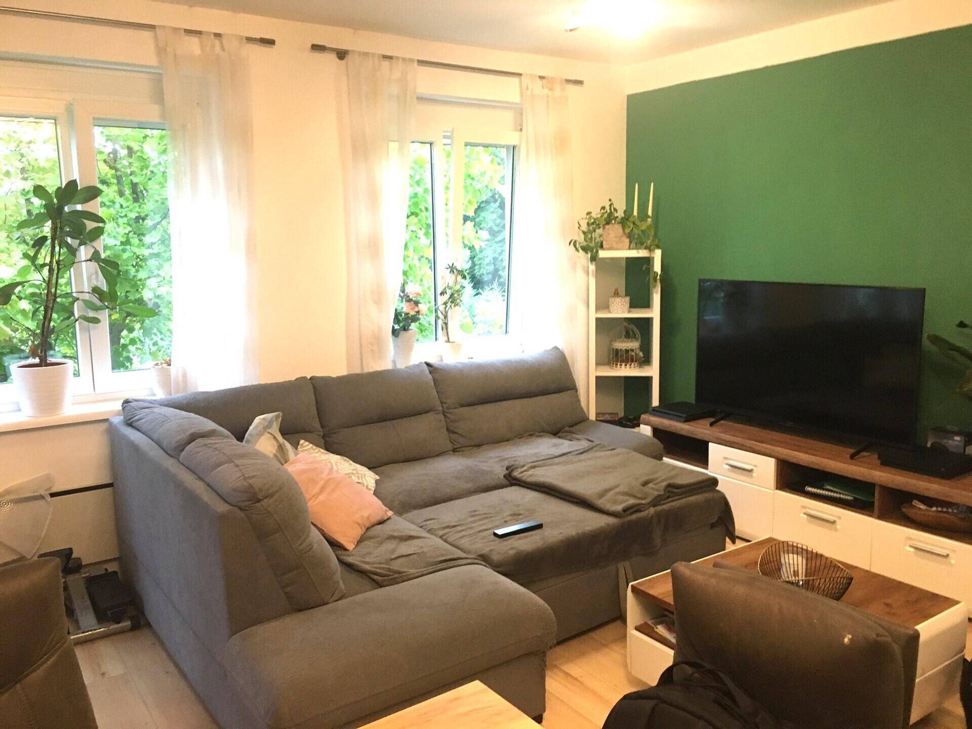 Helles Wohnzimmer mit großem Ecksofa, grüner Akzentwand und Blick ins Grüne.