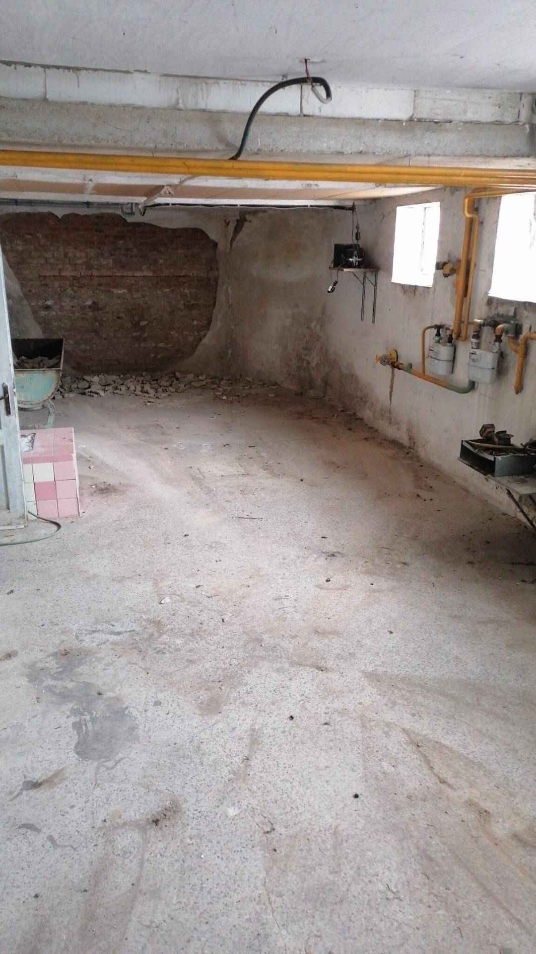 Unausgebauter Kellerraum mit Betonboden, sichtbaren Leitungen und Fenstern.