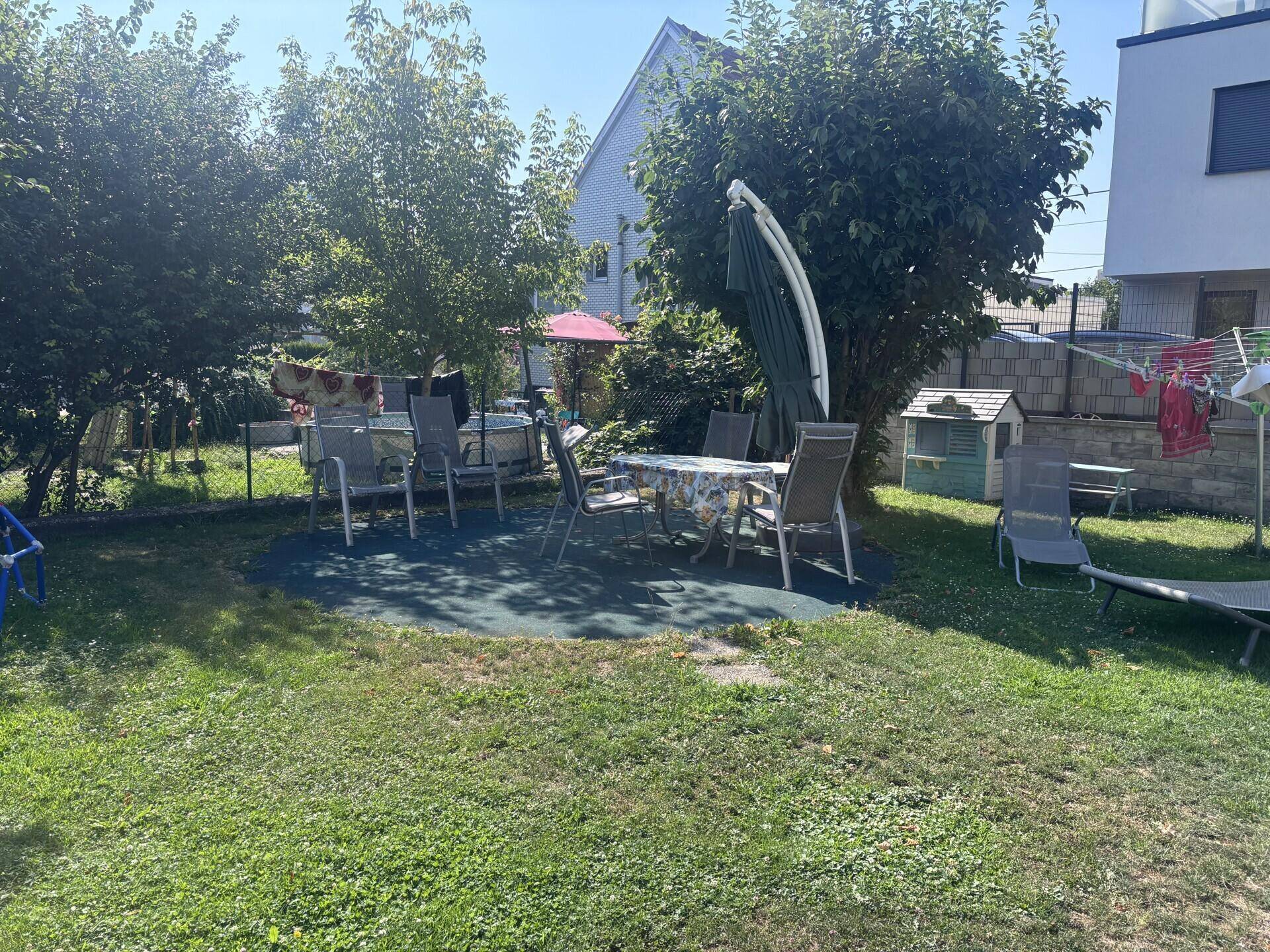 Sonniger Garten mit einer möblierten Sitzecke und Liegestühlen, umgeben von Rasen und Bäumen.