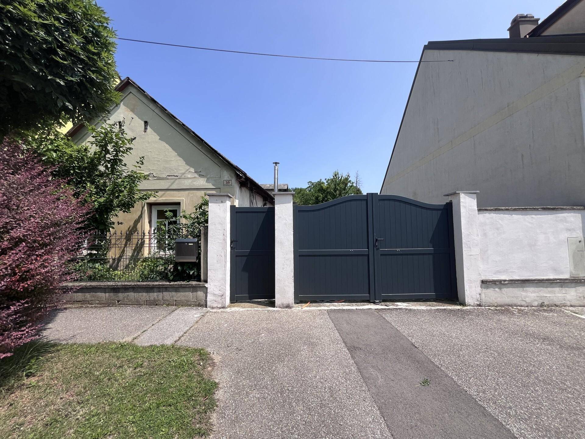 Hausansicht von der Straße mit dunklem Tor und Einfahrt unter blauem Himmel.