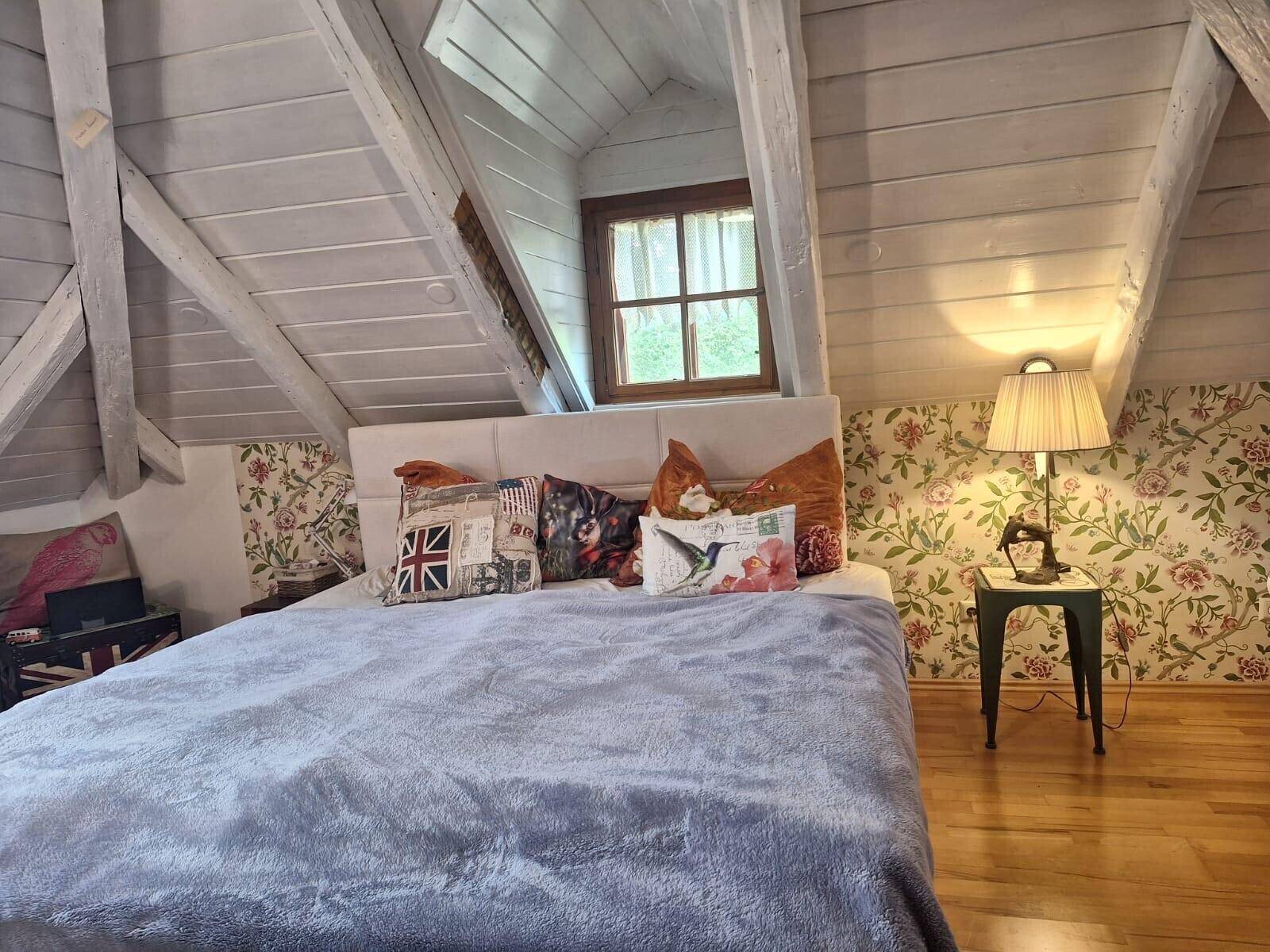 Gemütliches Schlafzimmer im Dachgeschoss mit Holzbalken, floraler Tapete und Holzboden.