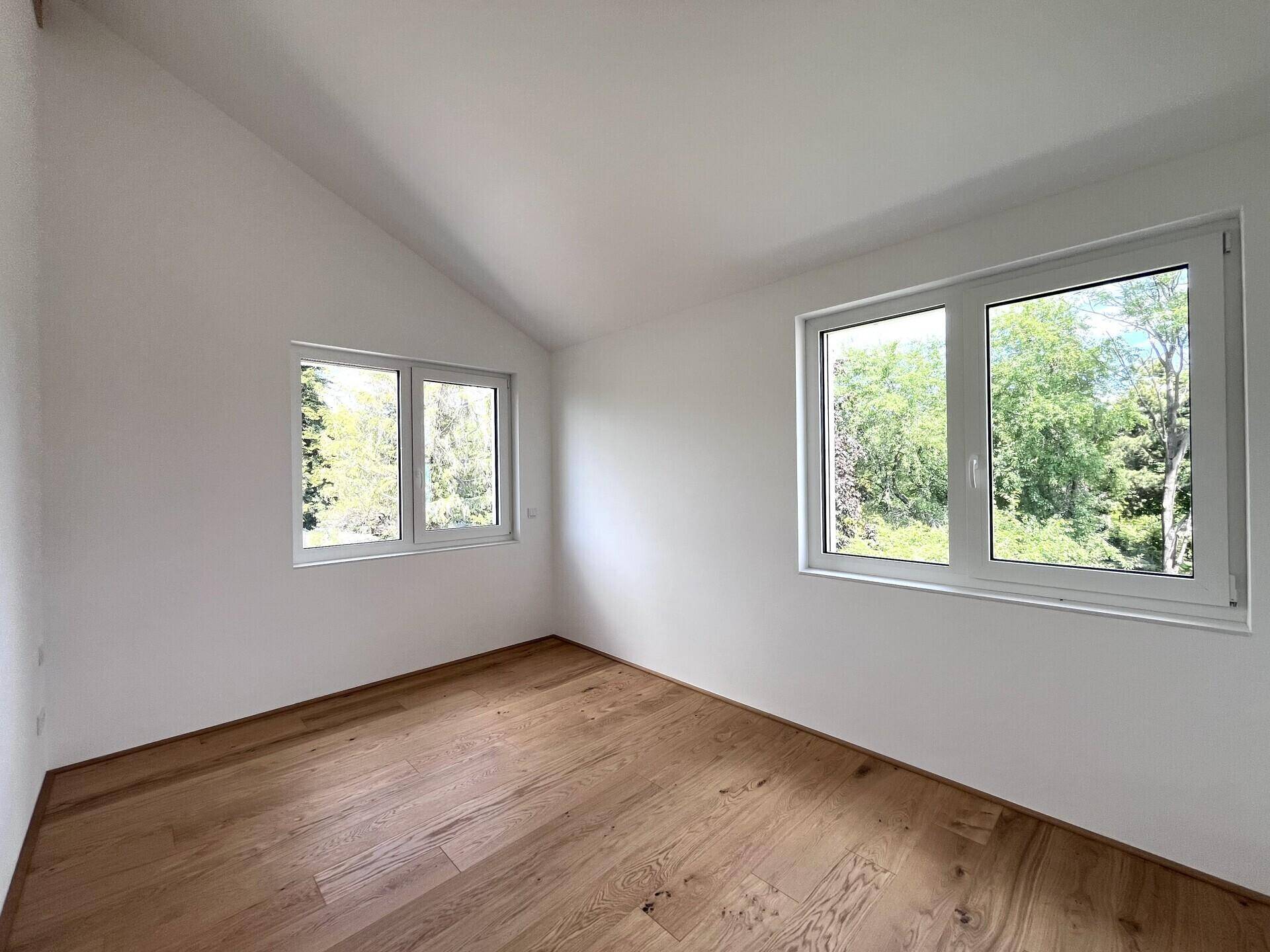 Helles Schlafzimmer mit Holzboden, Dachschräge und großen Fenstern mit Blick ins Grüne.