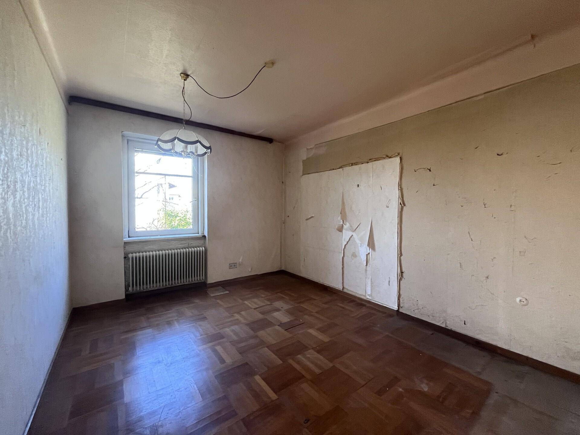 Zimmer mit Parkettboden, einem Fenster und beschädigter Tapete, ideal für eine Renovierung.
