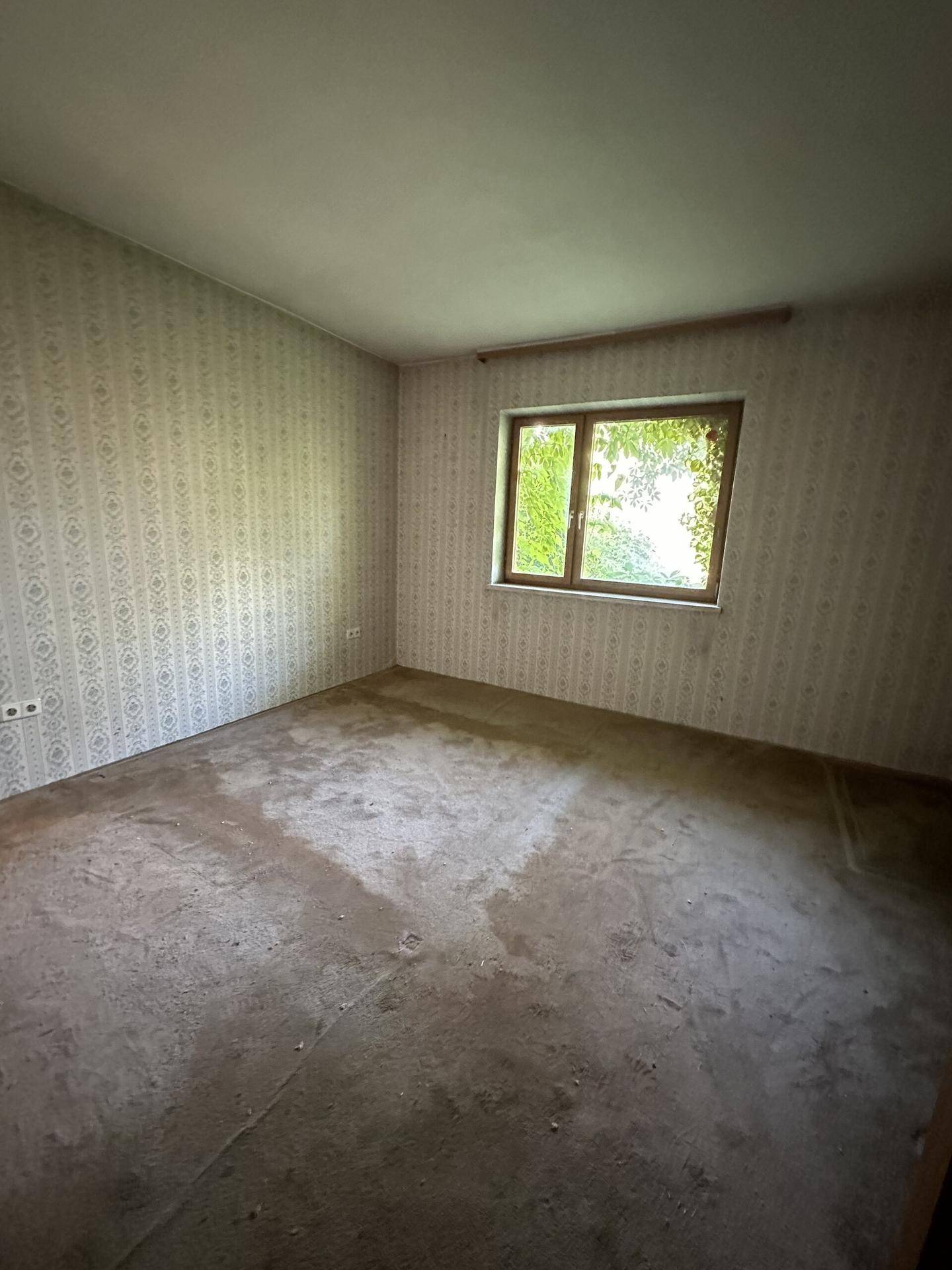 Leeres Schlafzimmer mit gemusterter Tapete und Fenster, das den Blick ins Grüne freigibt.