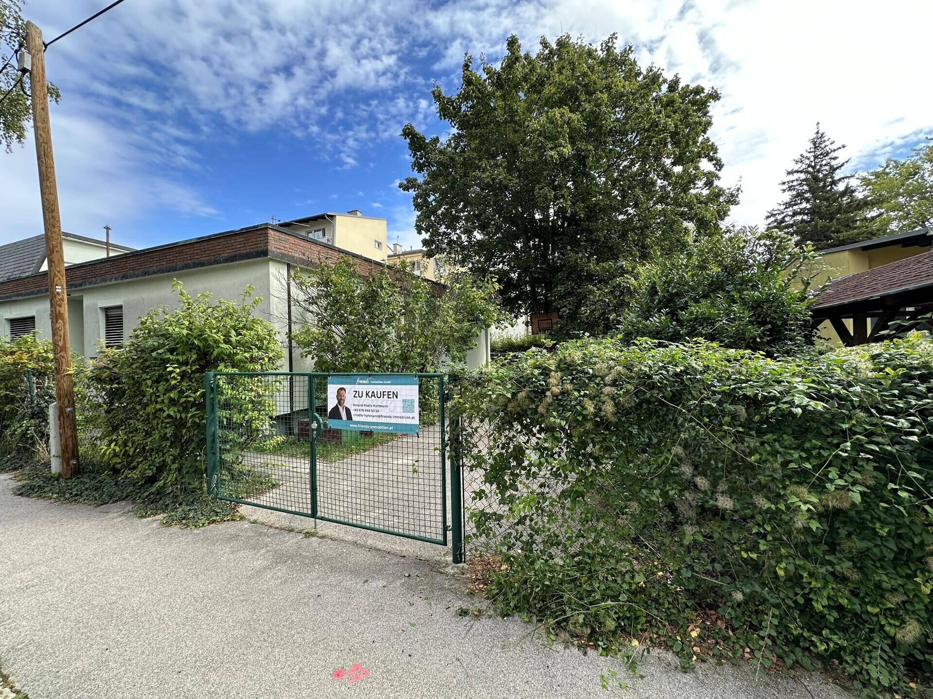 Gebäudeansicht von der Straße mit einem 'Zu Kaufen'-Schild und üppiger Vegetation.