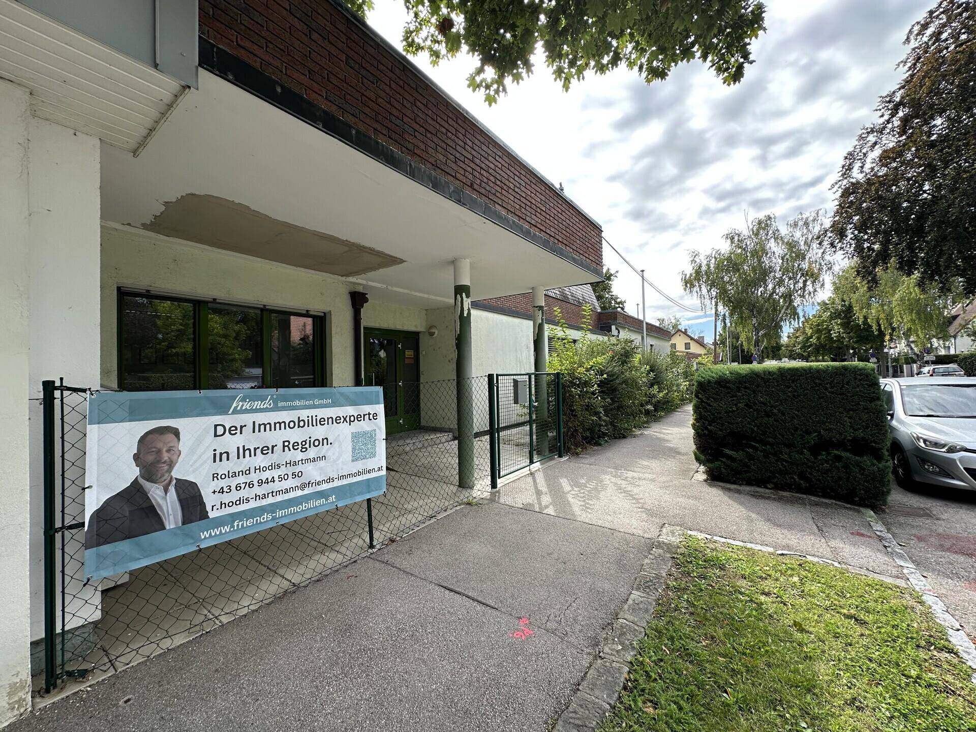 Eingangsbereich des Gebäudes mit einem Immobilienbanner und einem gepflegten Gehweg.