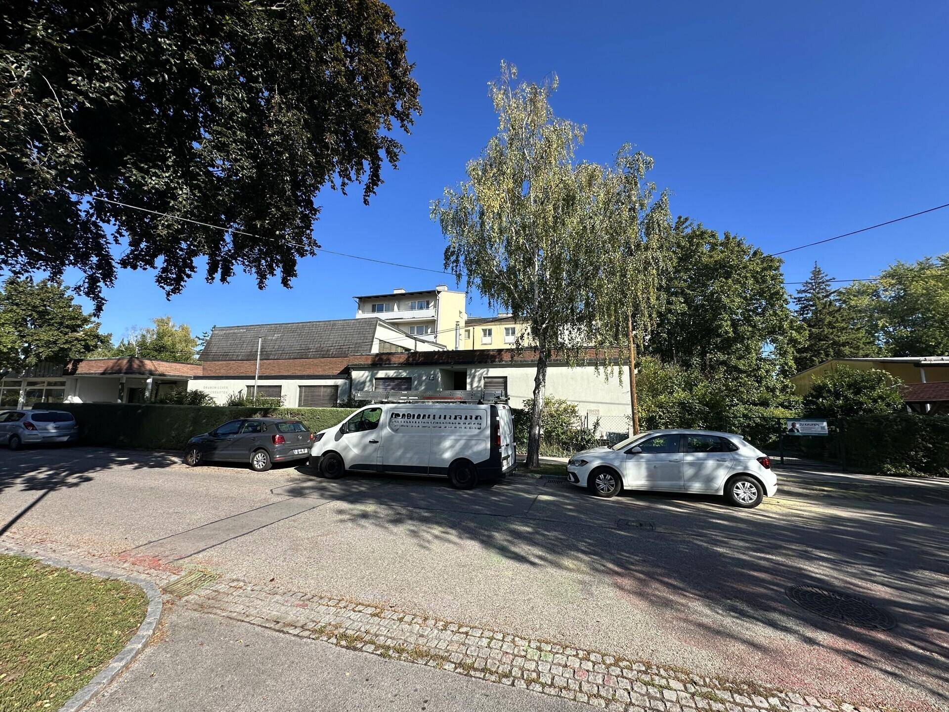 Außenansicht des Gebäudes mit parkenden Autos und Bäumen an einem klaren, sonnigen Tag.