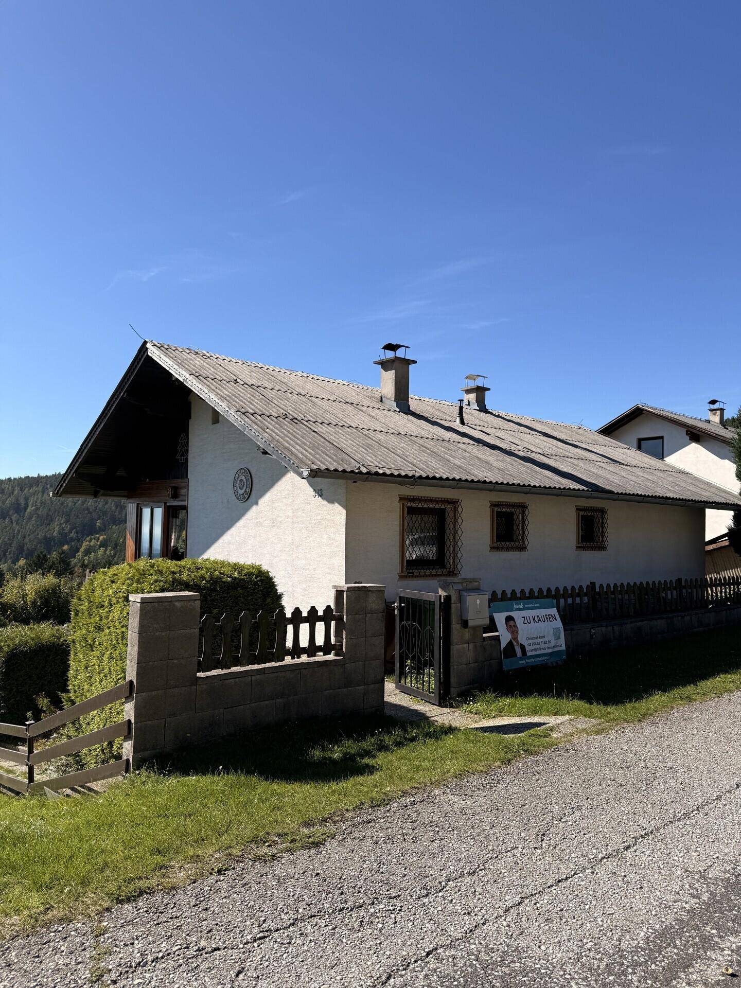 Vorderansicht des Hauses von der Straße aus, mit "Zu Kaufen"-Schild und traditionellem Dach.