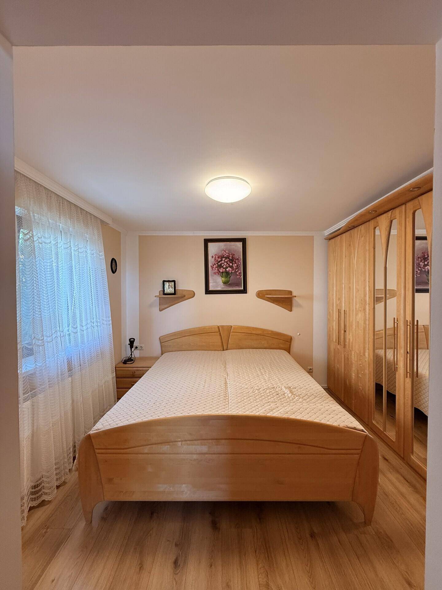 Helles Schlafzimmer mit Doppelbett, Holzschrank und Fenster mit Vorhängen.