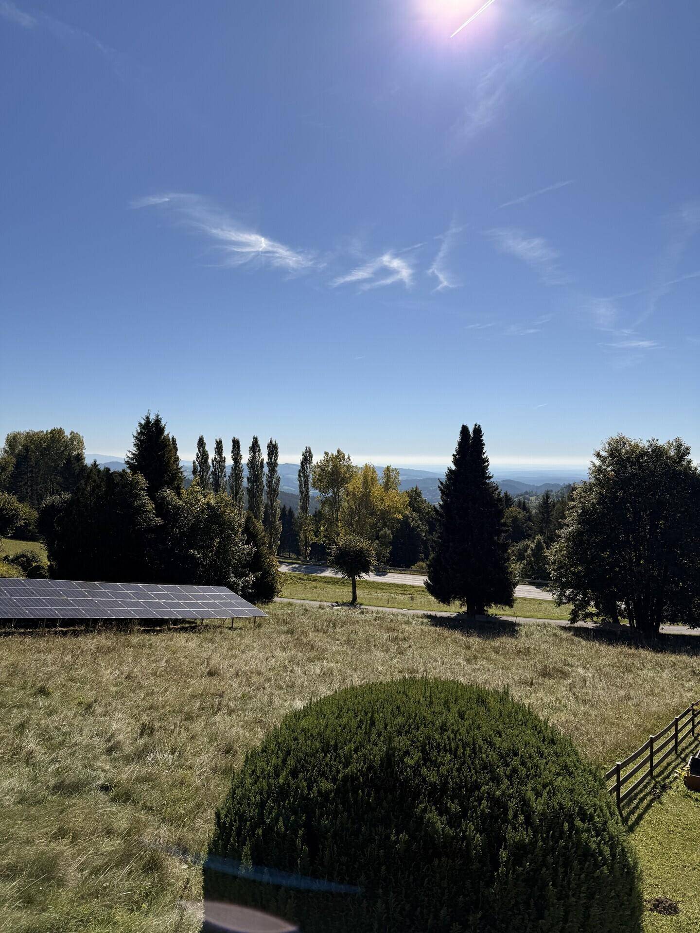 Weitläufige Landschaft mit Photovoltaikanlage, Bäumen und Bergen unter strahlend blauem Himmel.