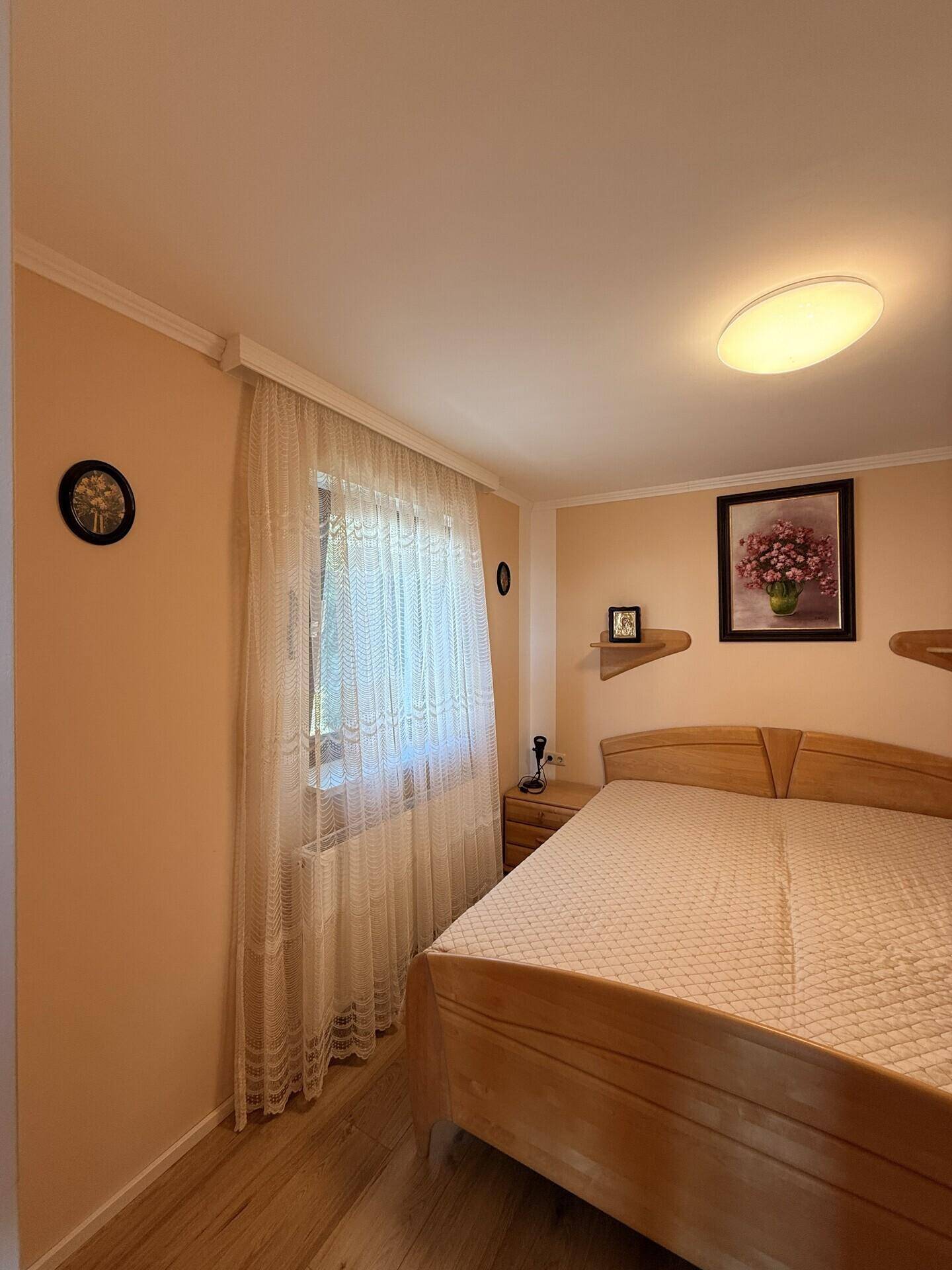 Gemütliches Schlafzimmer mit hellem Holzbett, Nachttisch und Fenster mit Gardine.
