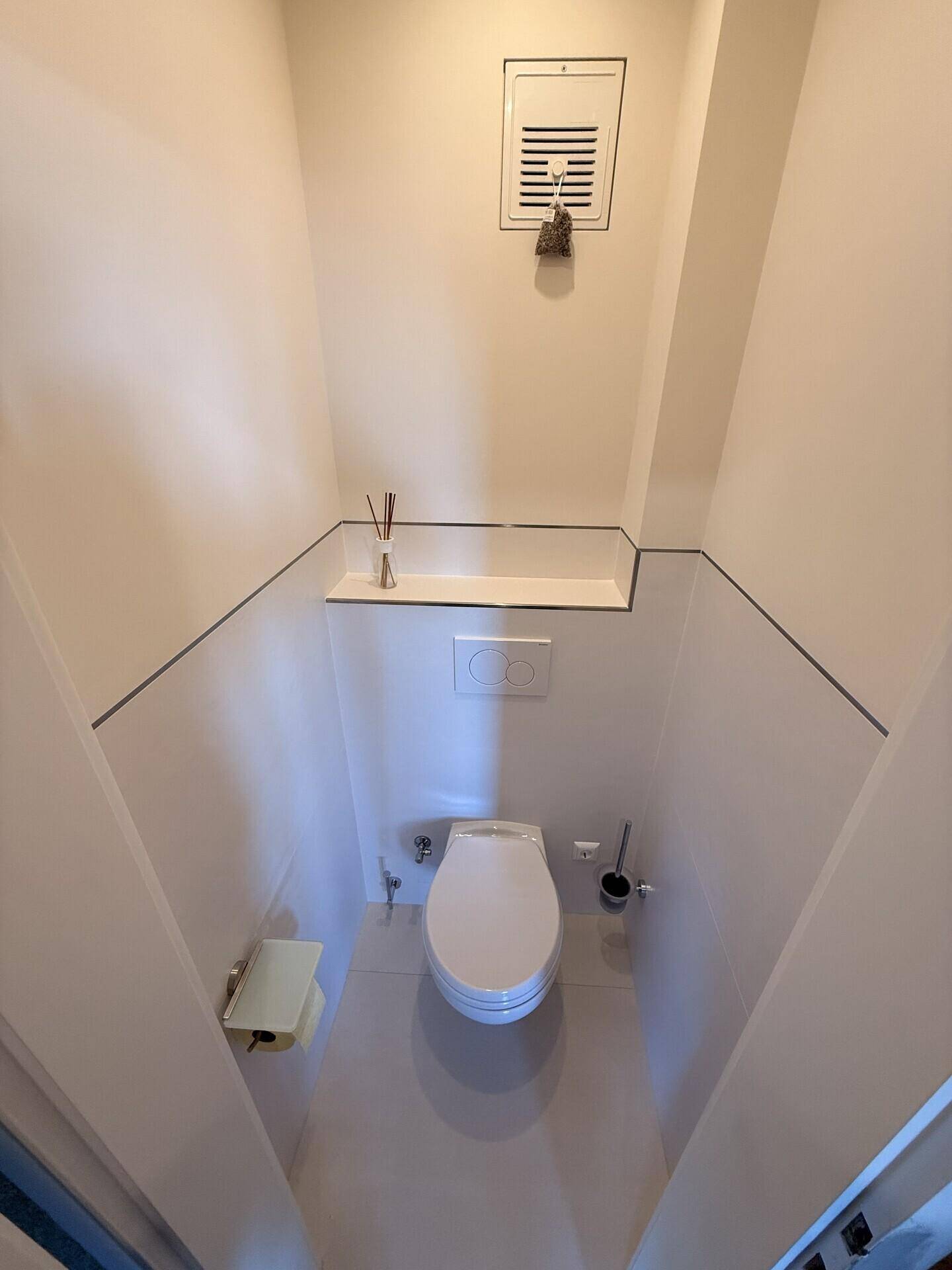 Sauberes Gäste-WC mit weißer Toilette, Wandspülkasten und Lüftungsgitter.