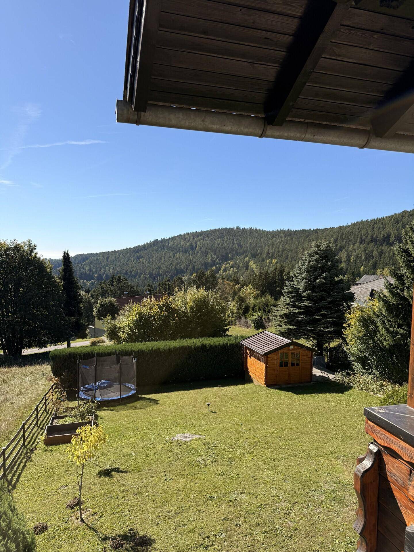 Blick vom Balkon auf den Garten mit Trampolin, Gartenhaus und grünen Hügeln im Hintergrund.