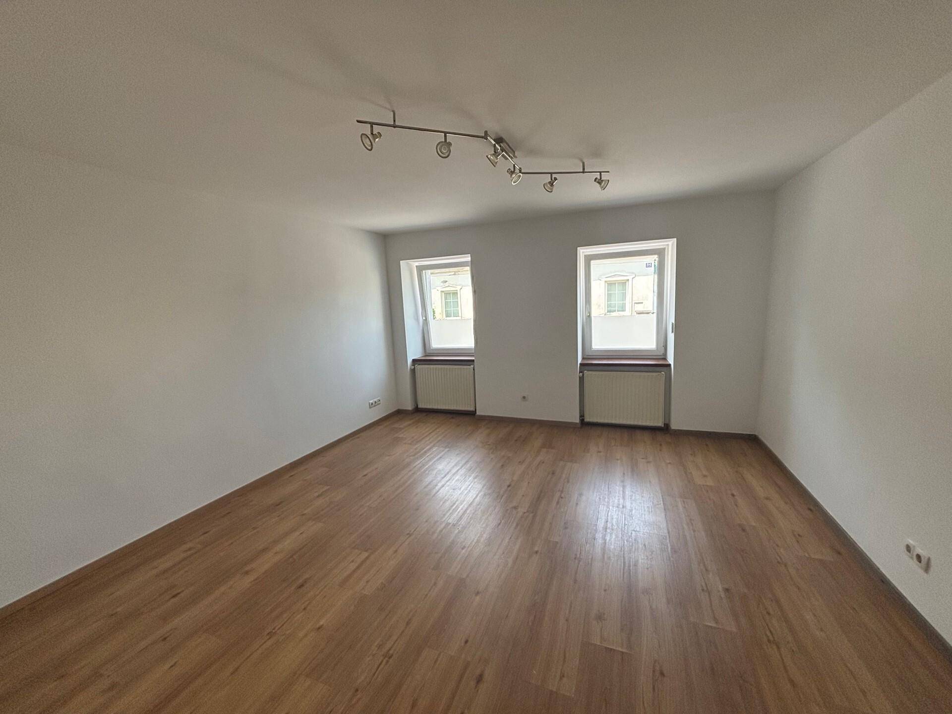 Helles, leeres Wohnzimmer mit Laminatboden und zwei Fenstern, bereit zur Einrichtung.