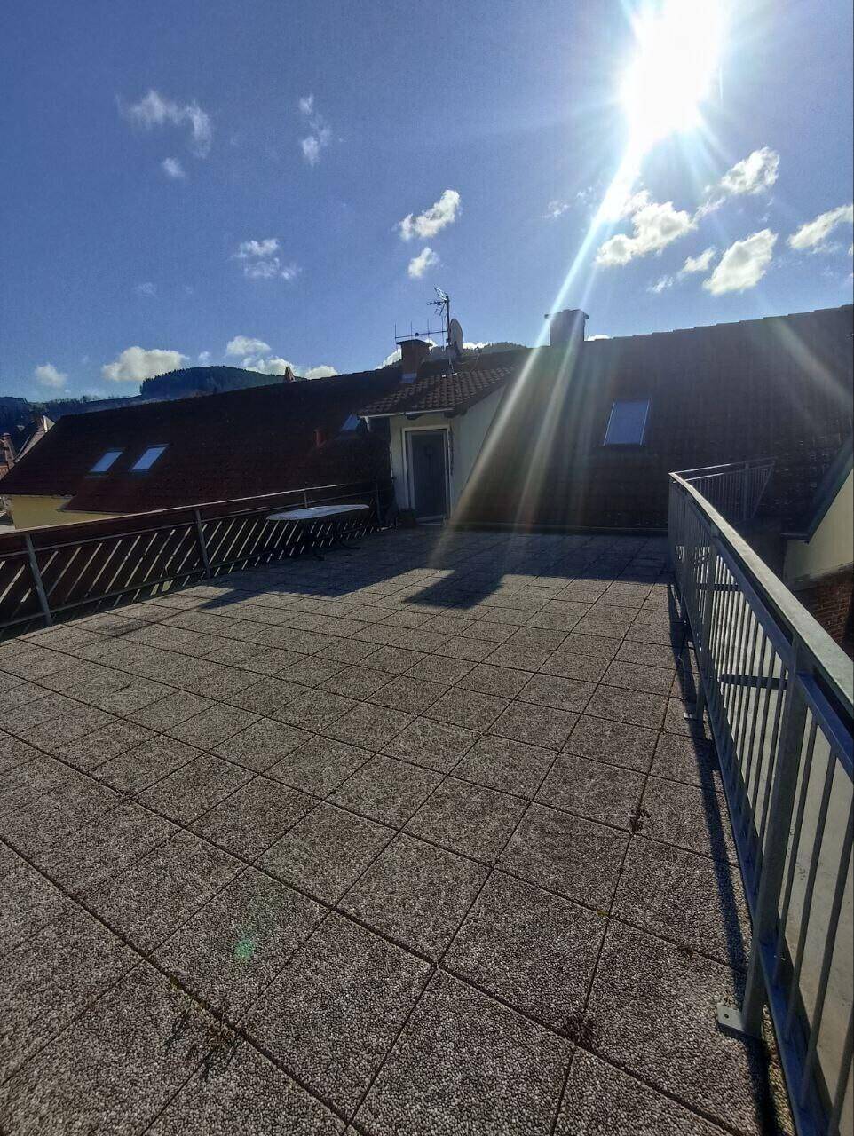 Große Dachterrasse mit Fliesenboden und Metallgeländer unter strahlendem Sonnenschein.