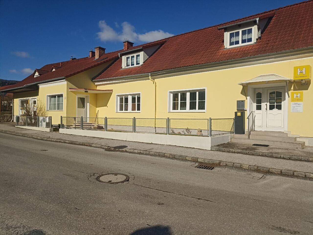 Ansicht der gelben Gebäudefassade mit Eingangsbereich und Fenstern zur Straße hin.