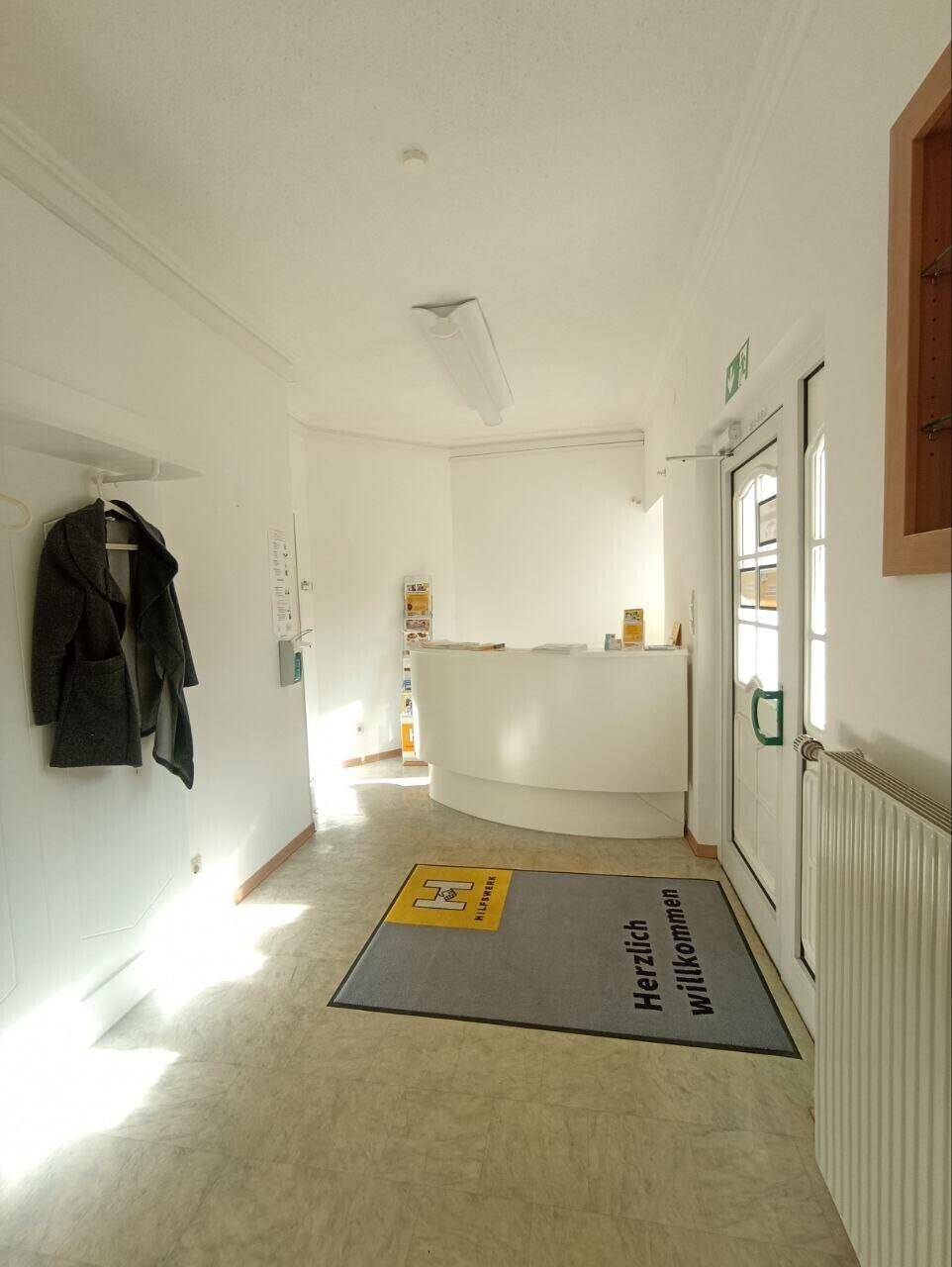 Heller Empfangsbereich mit Rezeptionstheke, Garderobe und Willkommensmatte.