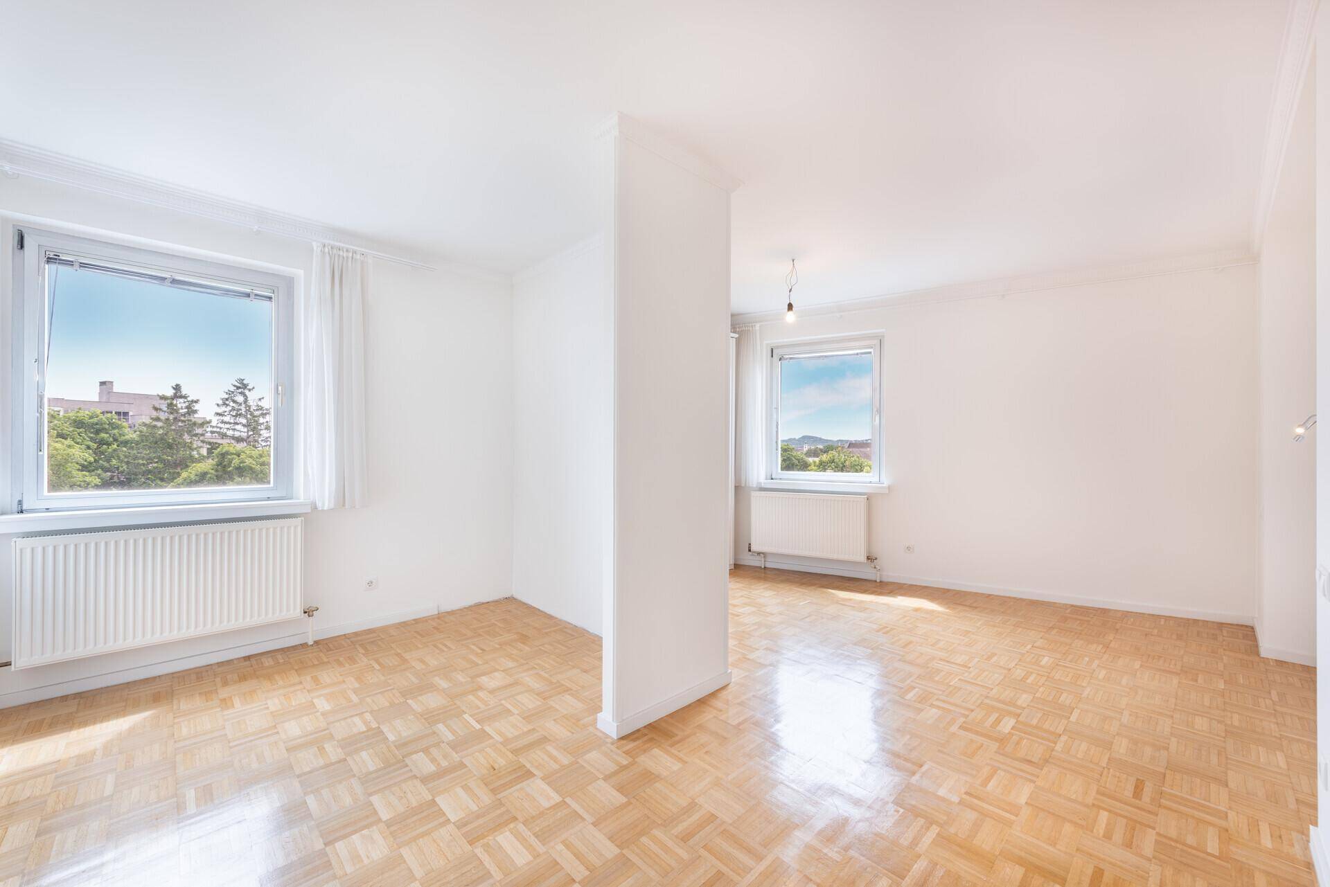 Leeres Zimmer mit hellem Parkettboden und zwei Fenstern, die viel Tageslicht hereinlassen.