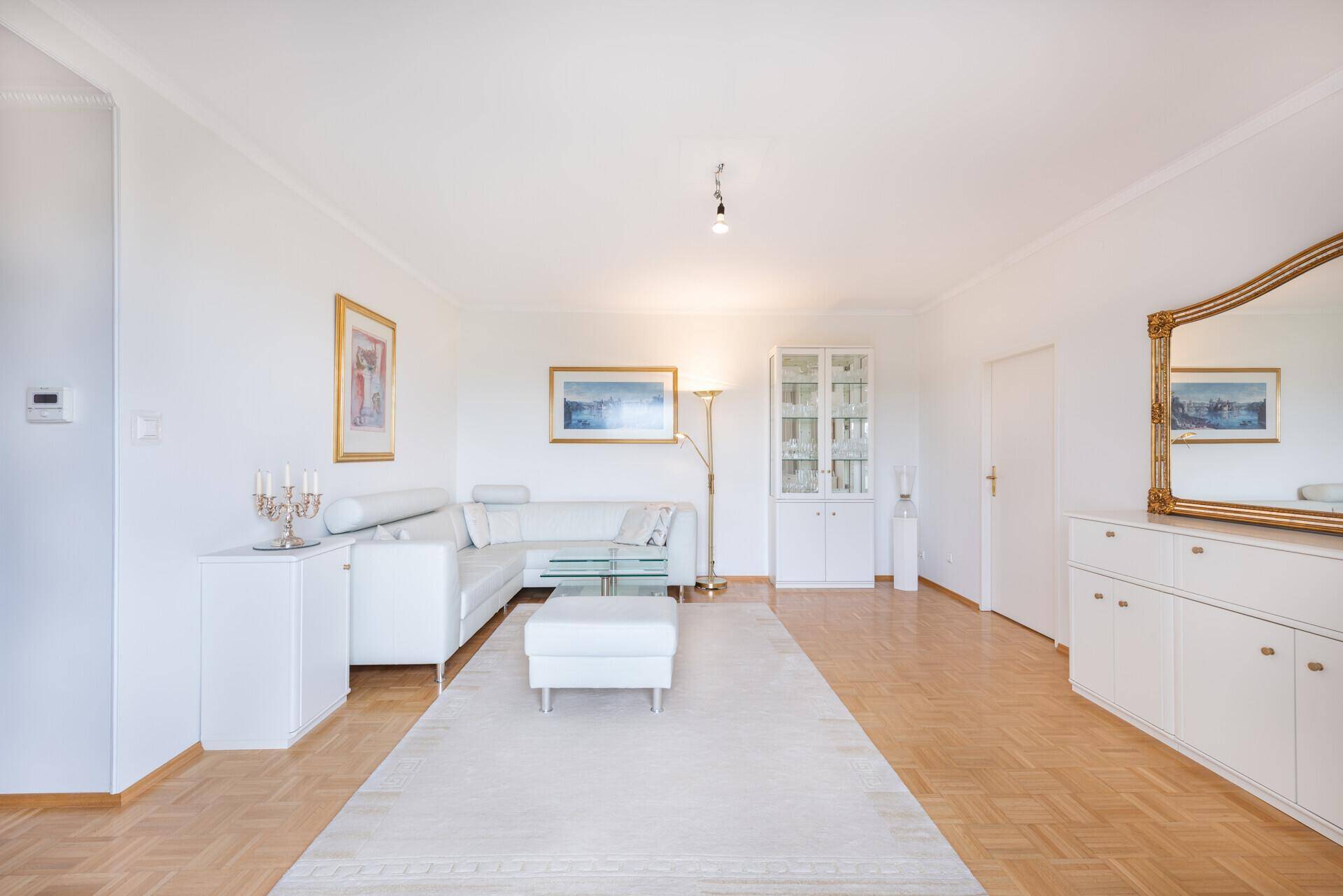 Geräumiges Wohnzimmer mit hellem Parkettboden, weißer Einrichtung und einem großen Wandspiegel.