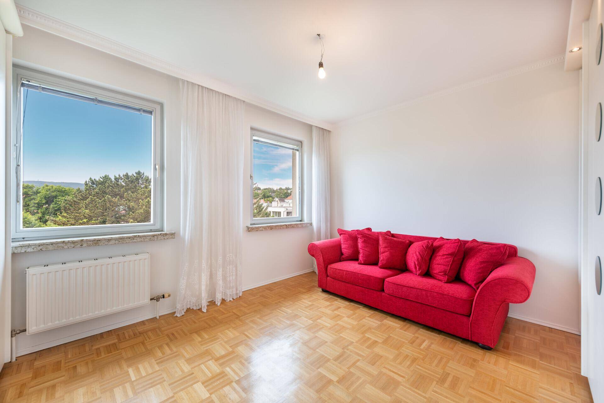 Helles Zimmer mit Parkettboden, zwei Fenstern und einem auffälligen roten Sofa.