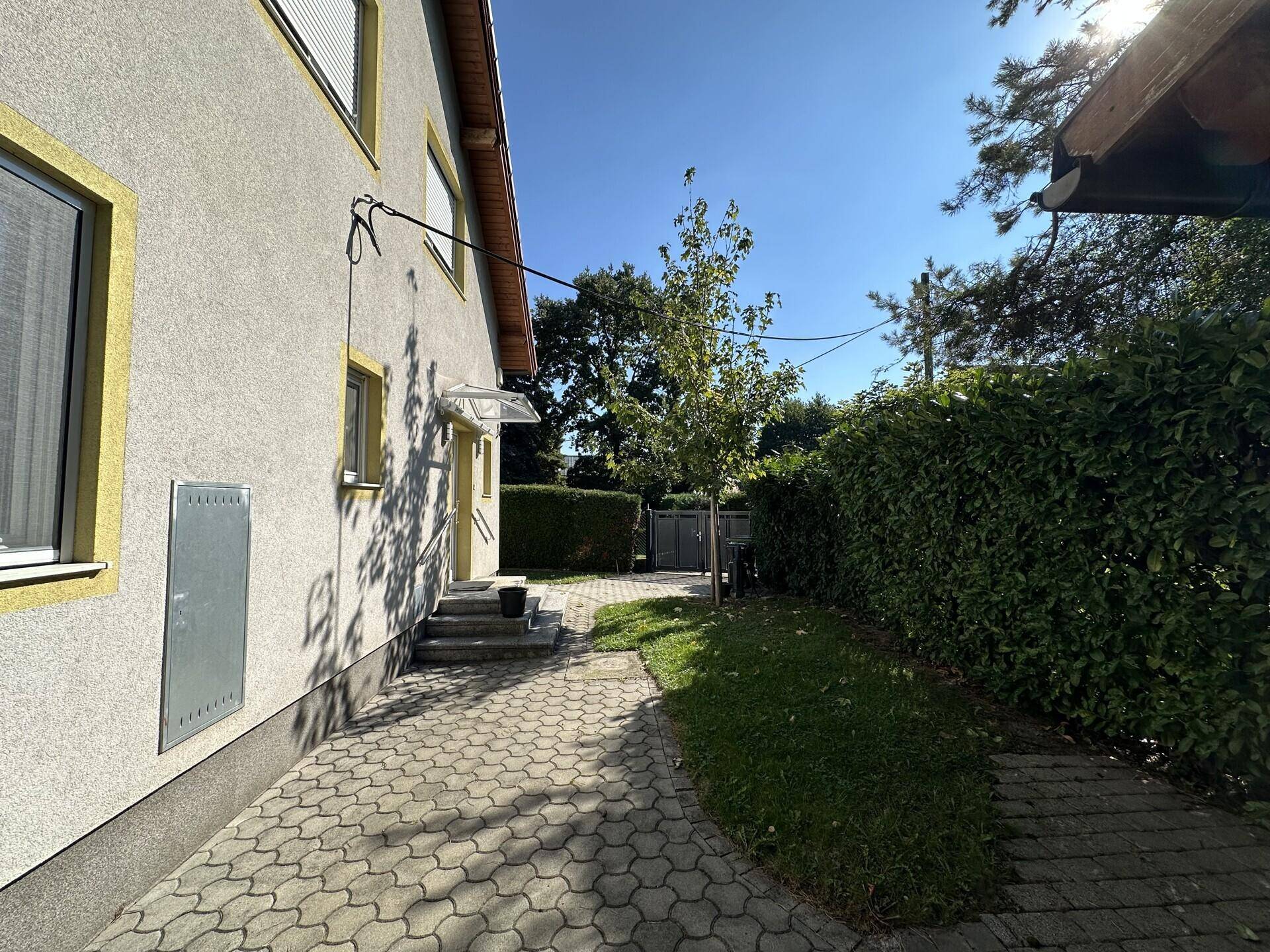 Seitenansicht des Hauses mit gepflastertem Weg, umgeben von hohen Hecken und Bäumen.