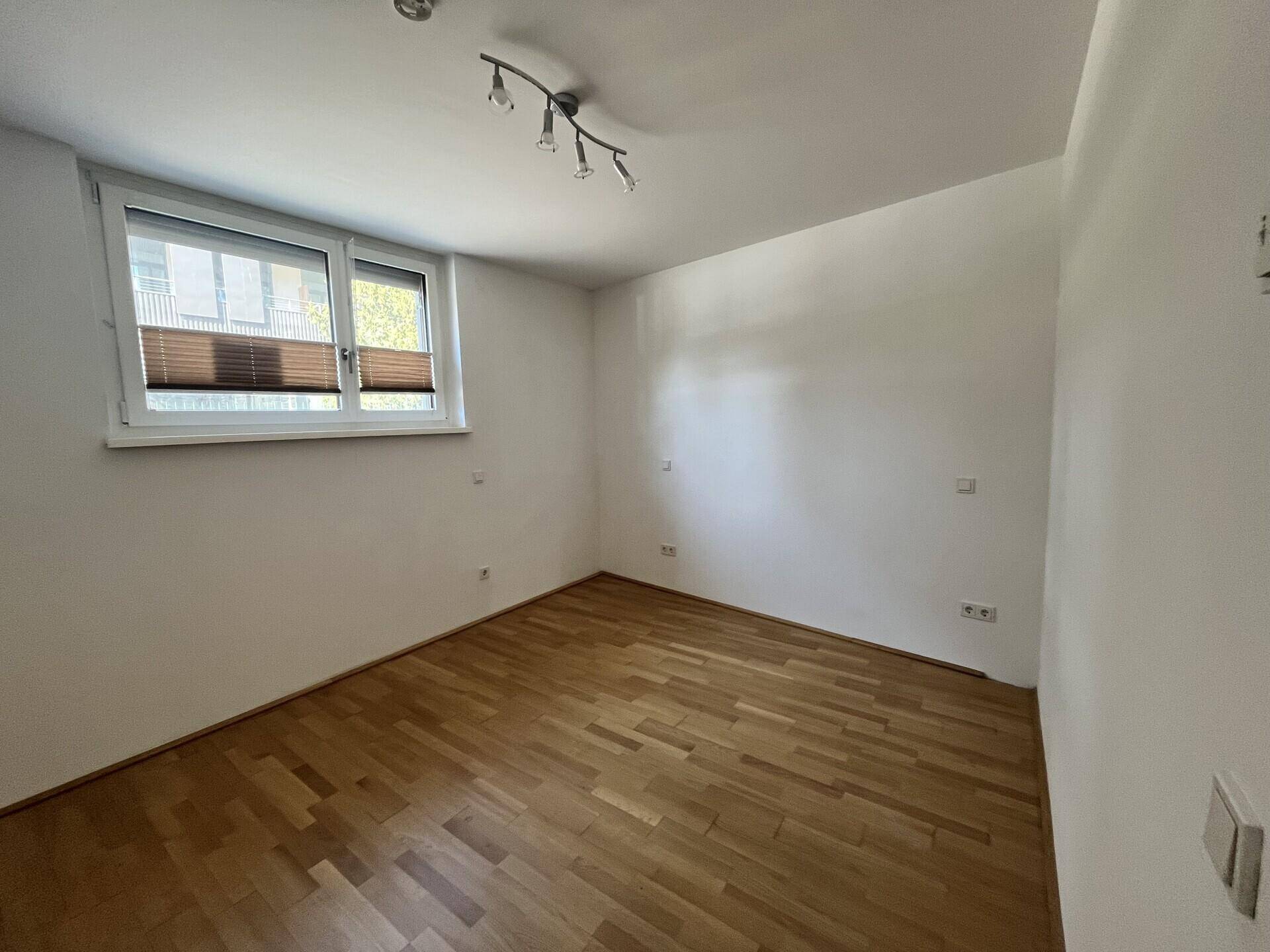 Schlafzimmer mit Parkettboden, weißen Wänden und Fenster mit Jalousien.
