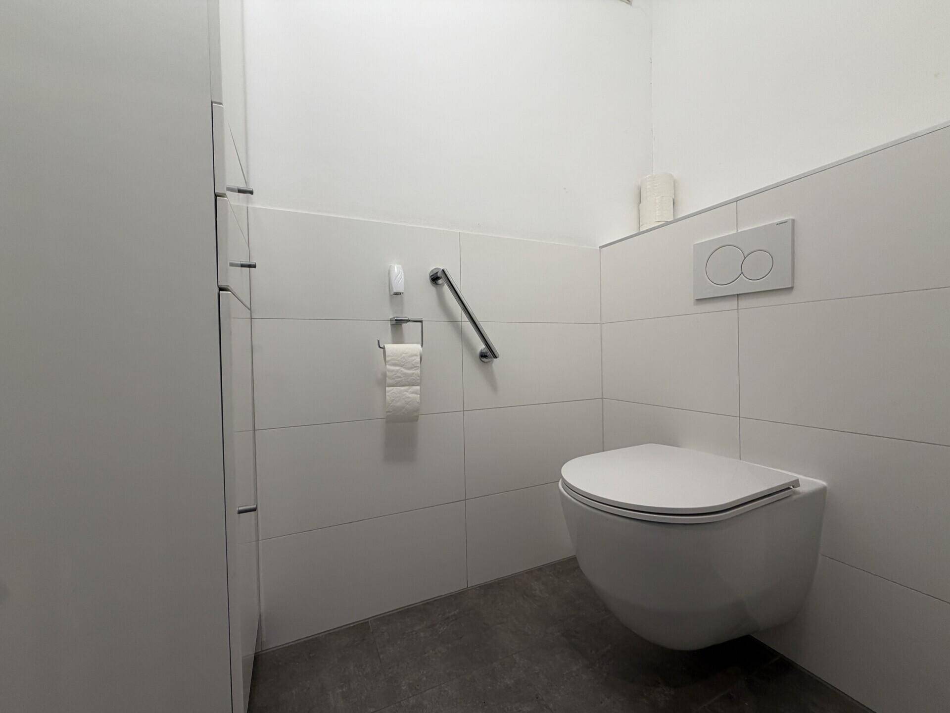 Modernes Gäste-WC mit wandhängender Toilette, weißen Fliesen und Haltegriff.