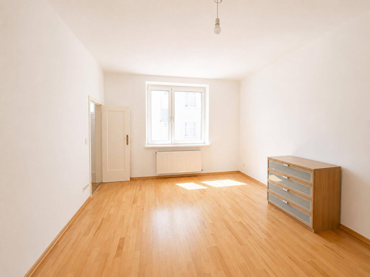 Helles Schlafzimmer mit Parkettboden, Fenster und Kommode.