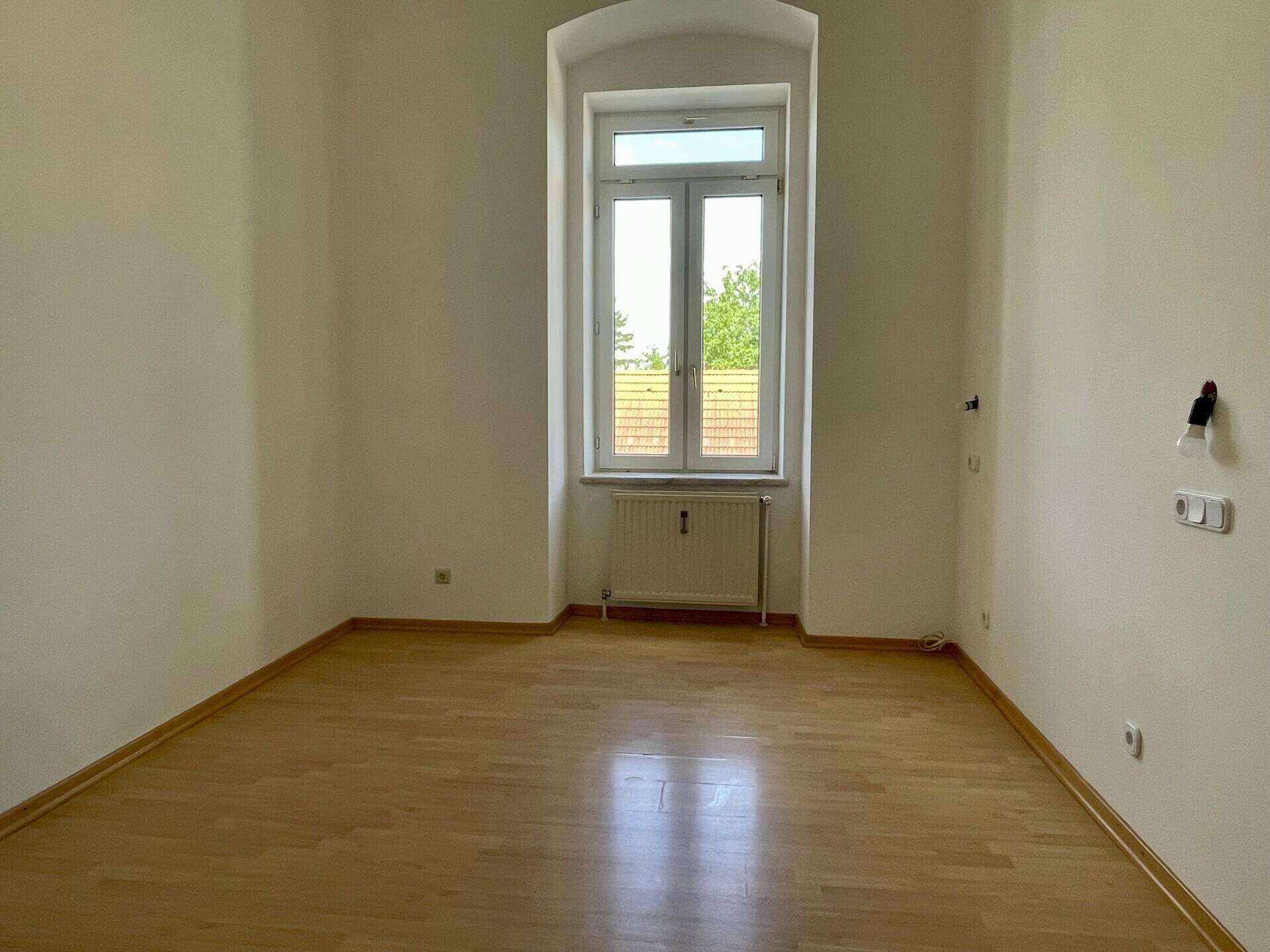 Helles Zimmer mit Holzboden und großem Fenster, das auf die Einrichtung wartet.