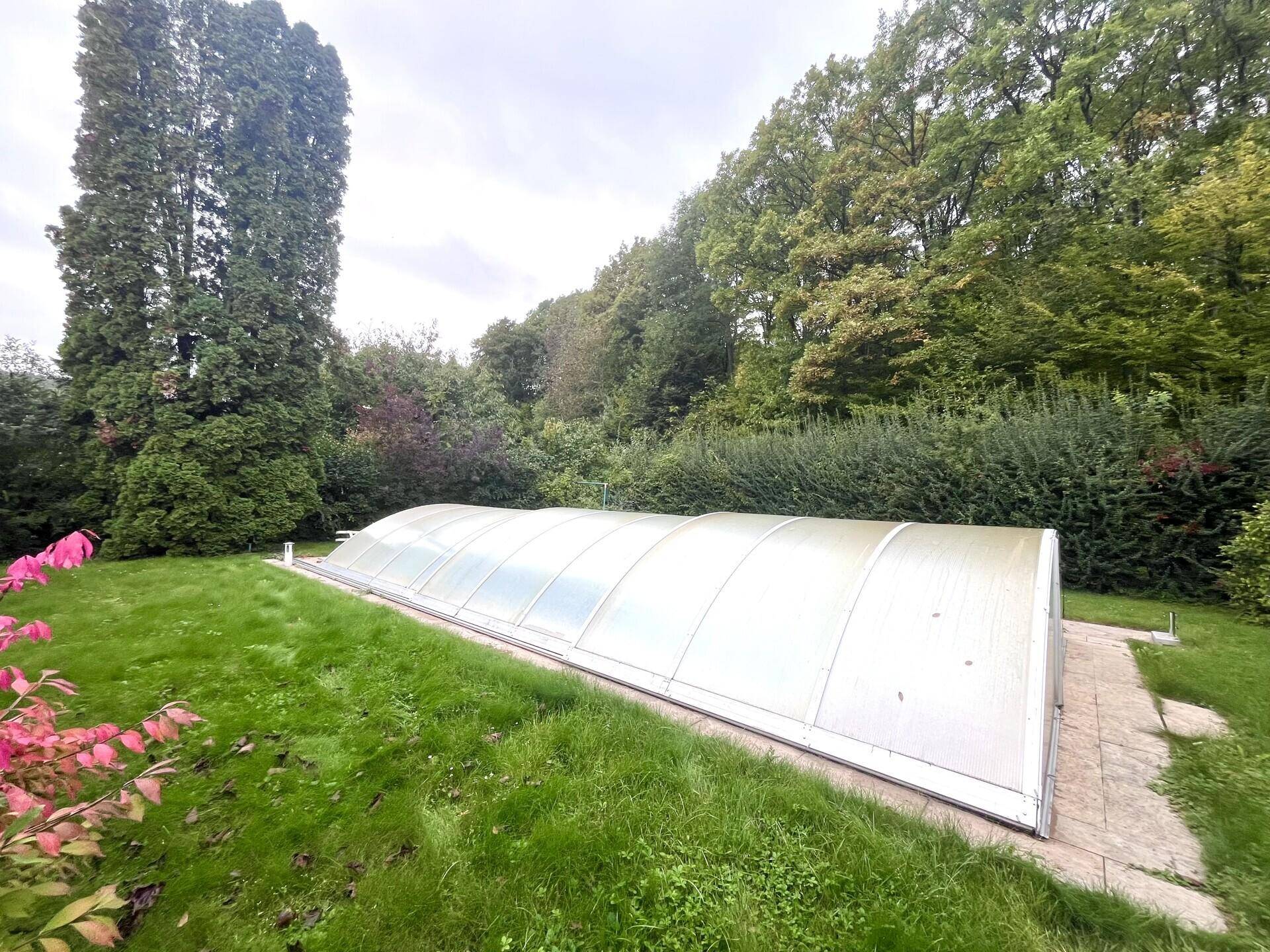 Weitläufiger Garten mit Rasen, Bäumen und einem überdachten Swimmingpool im Hintergrund.