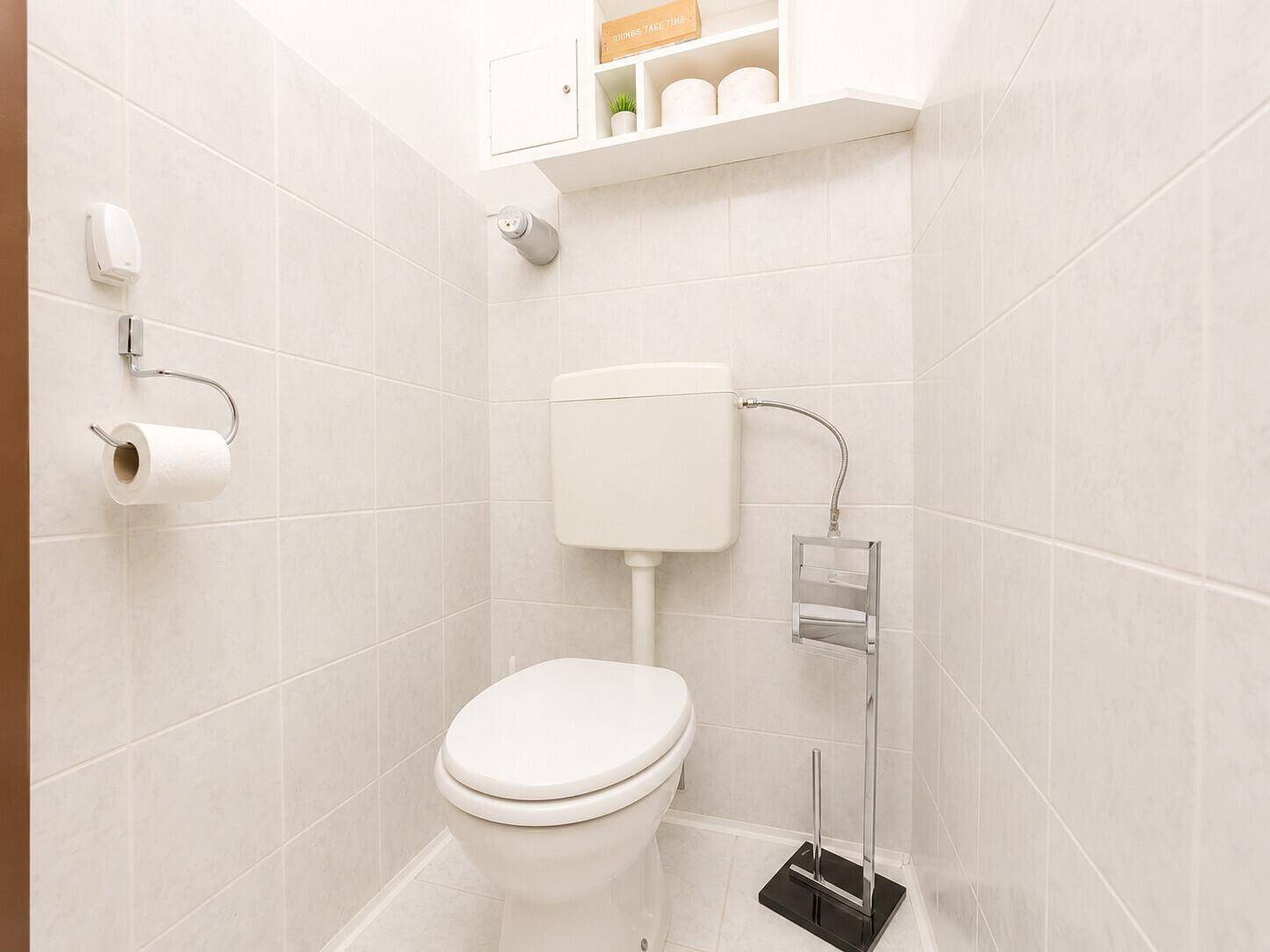 Separates WC mit weiß gefliesten Wänden und Boden, ausgestattet mit Toilette und Regal.