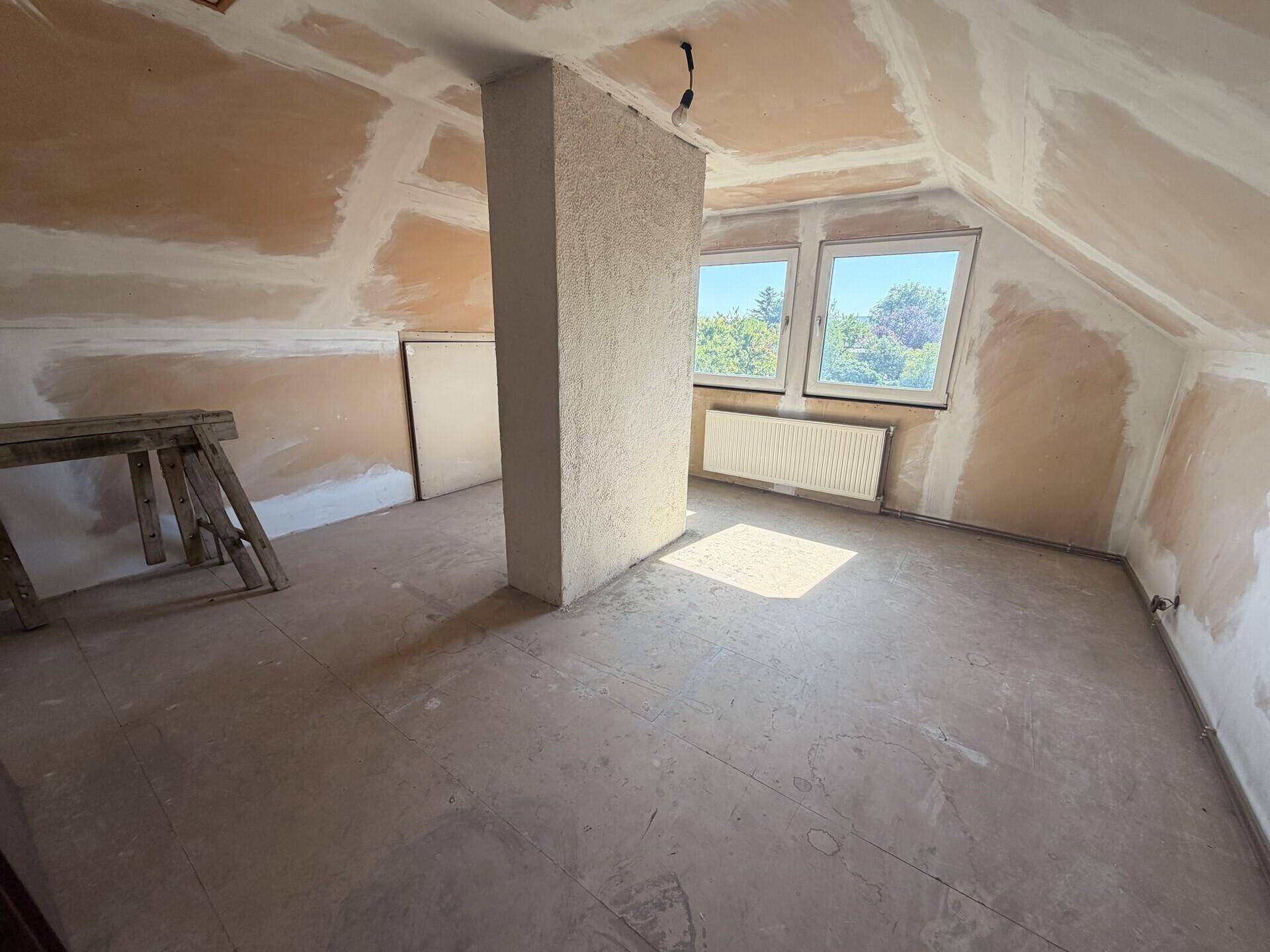 Helles Dachgeschosszimmer im Rohbau mit zwei Fenstern und Heizkörper.