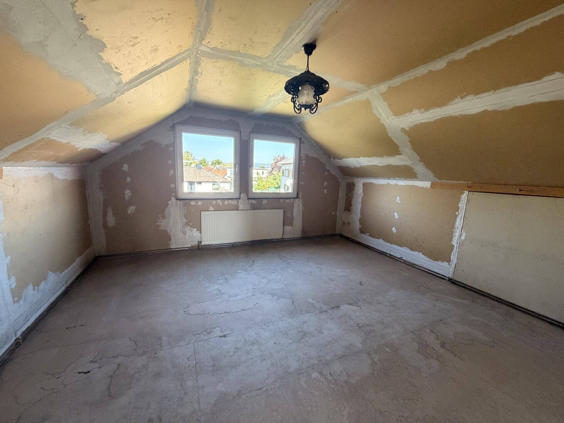 Dachgeschosszimmer im Rohbau mit Fenster und alter Deckenleuchte.