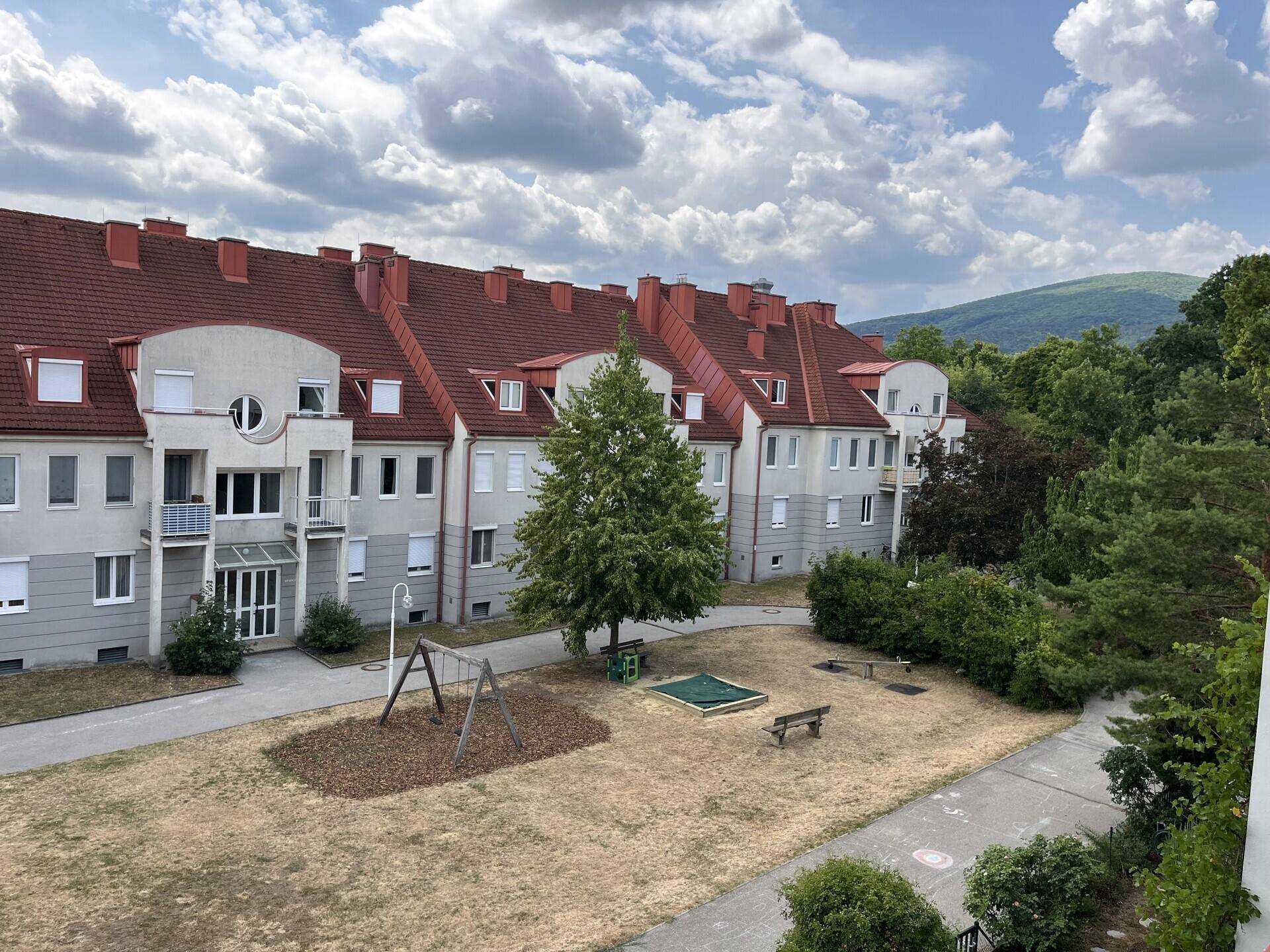 Wohnanlage mit rotem Ziegeldach, Innenhof und Kinderspielplatz unter blauem Himmel.