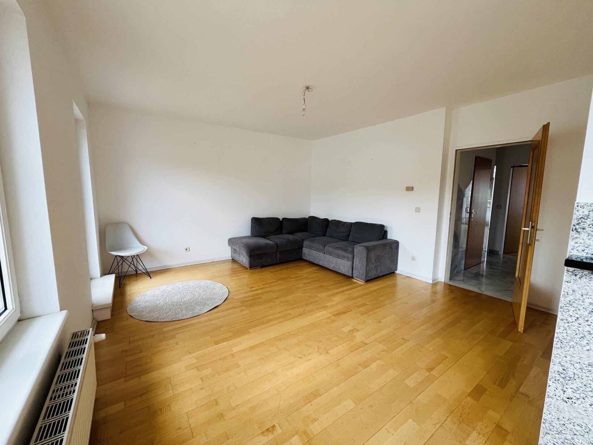 Wohnzimmer mit Parkettboden und grauer Eckcouch, Blick auf den Flur und die Küche.