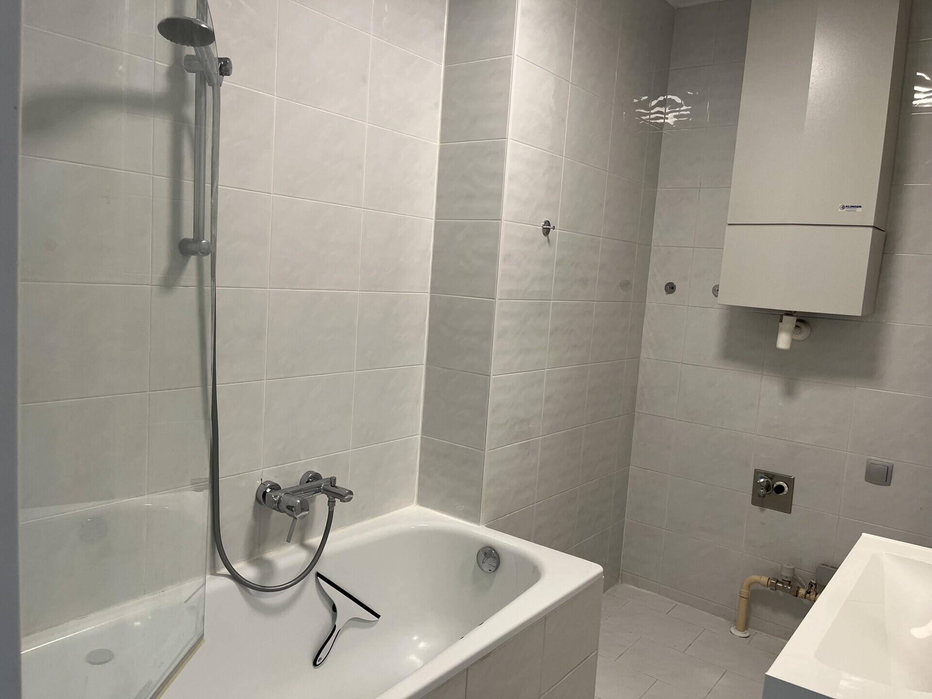 Badezimmer mit Badewanne, Duscharmatur und hellen Fliesen, Heiztherme an der Wand.