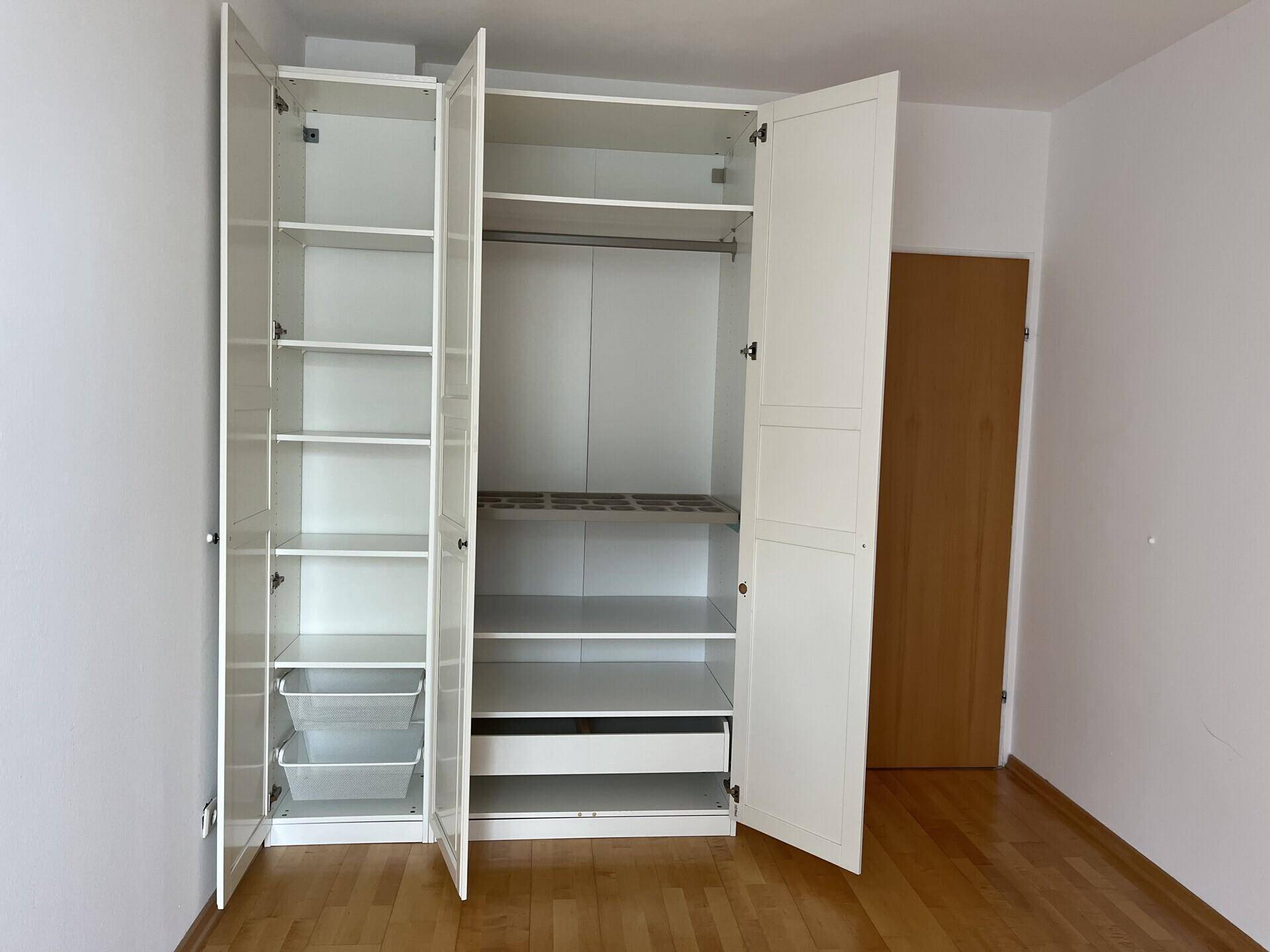 Weißer Kleiderschrank mit offenen Türen und verschiedenen Ablagefächern in einem hellen Raum.