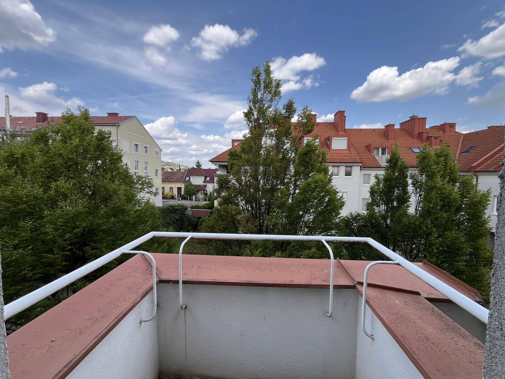 Blick von einem Balkon auf Wohngebäude mit roten Dächern und üppigem Grün unter blauem Himmel.