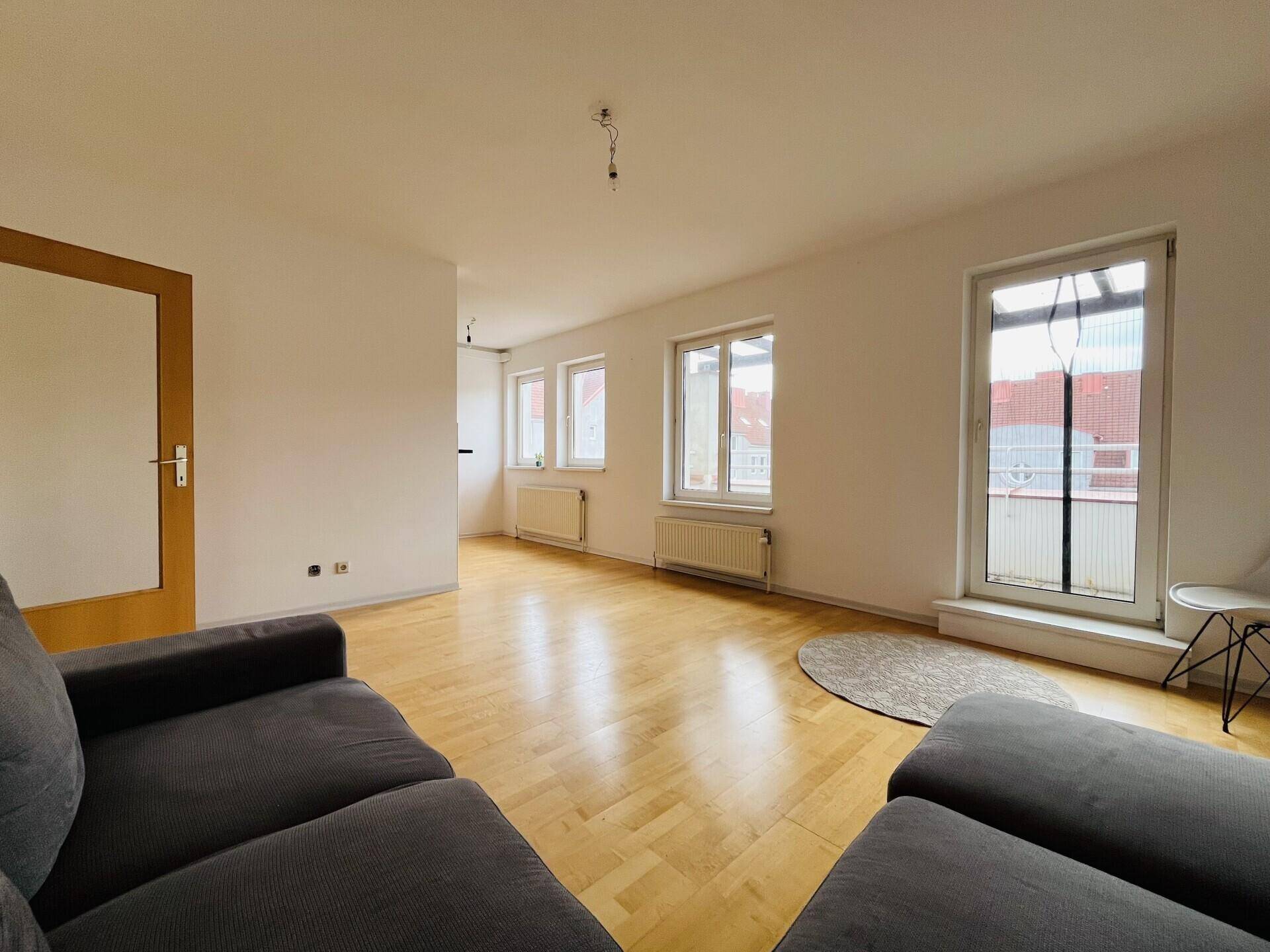 Geräumiges Wohnzimmer mit hellem Parkettboden, großen Fenstern und Zugang zum Balkon.