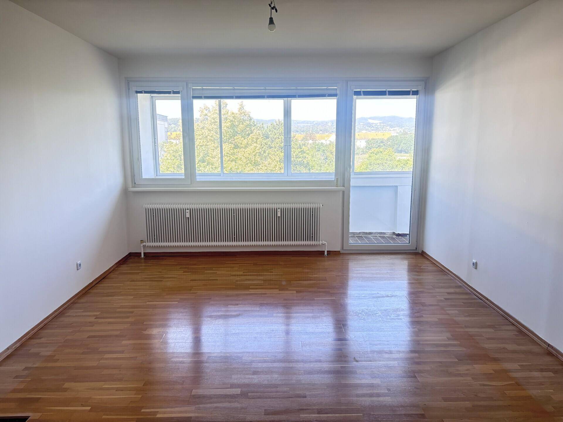 Helles, leeres Zimmer mit Parkettboden, großen Fenstern und Zugang zu einem kleinen Balkon.
